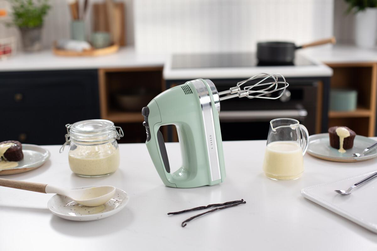 Ein mintgrüner Handmixer auf einer weißen Arbeitsplatte, mit Backzubehör.