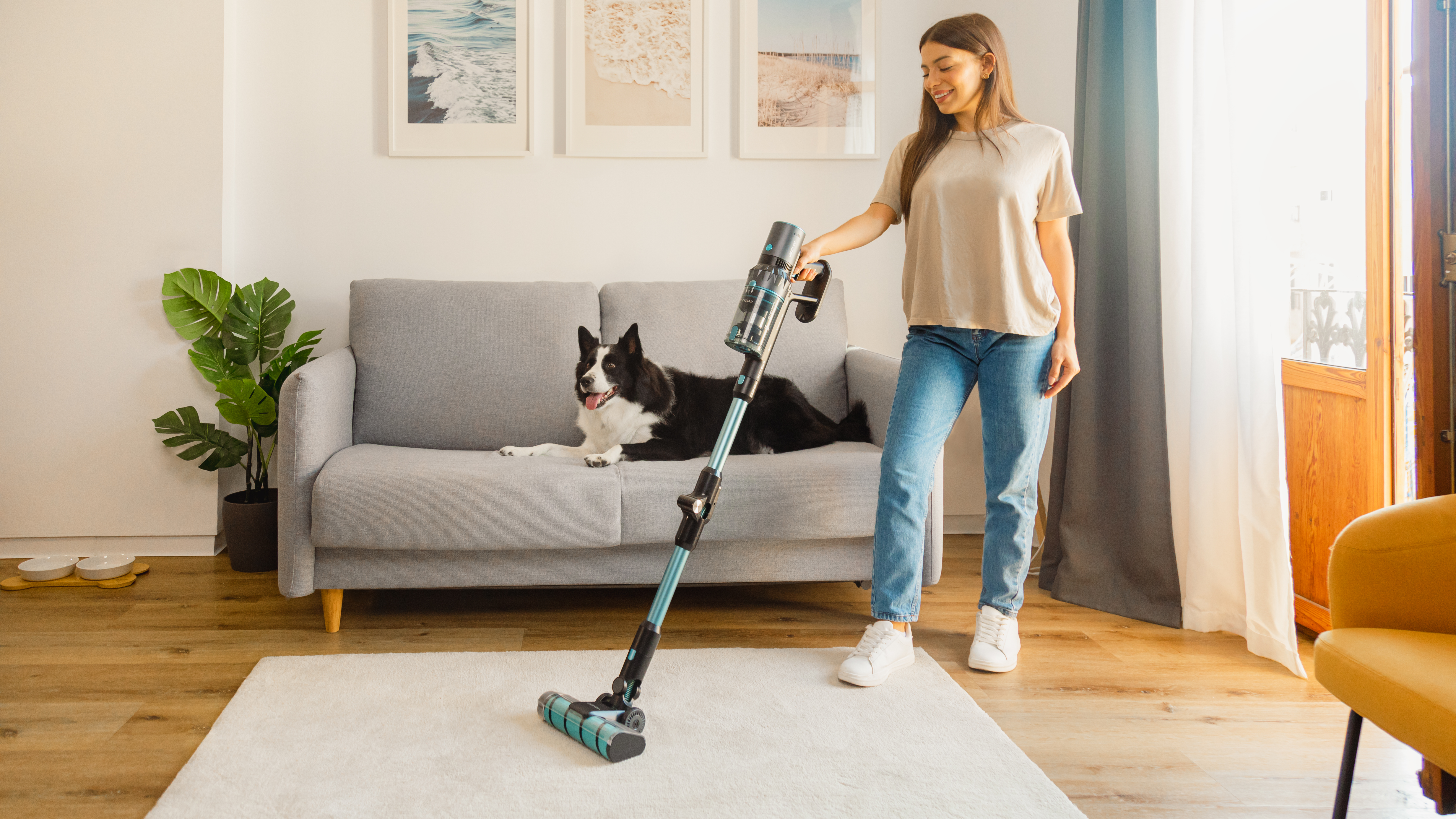 Una mujer aspira una alfombra con un perro en un sofá.