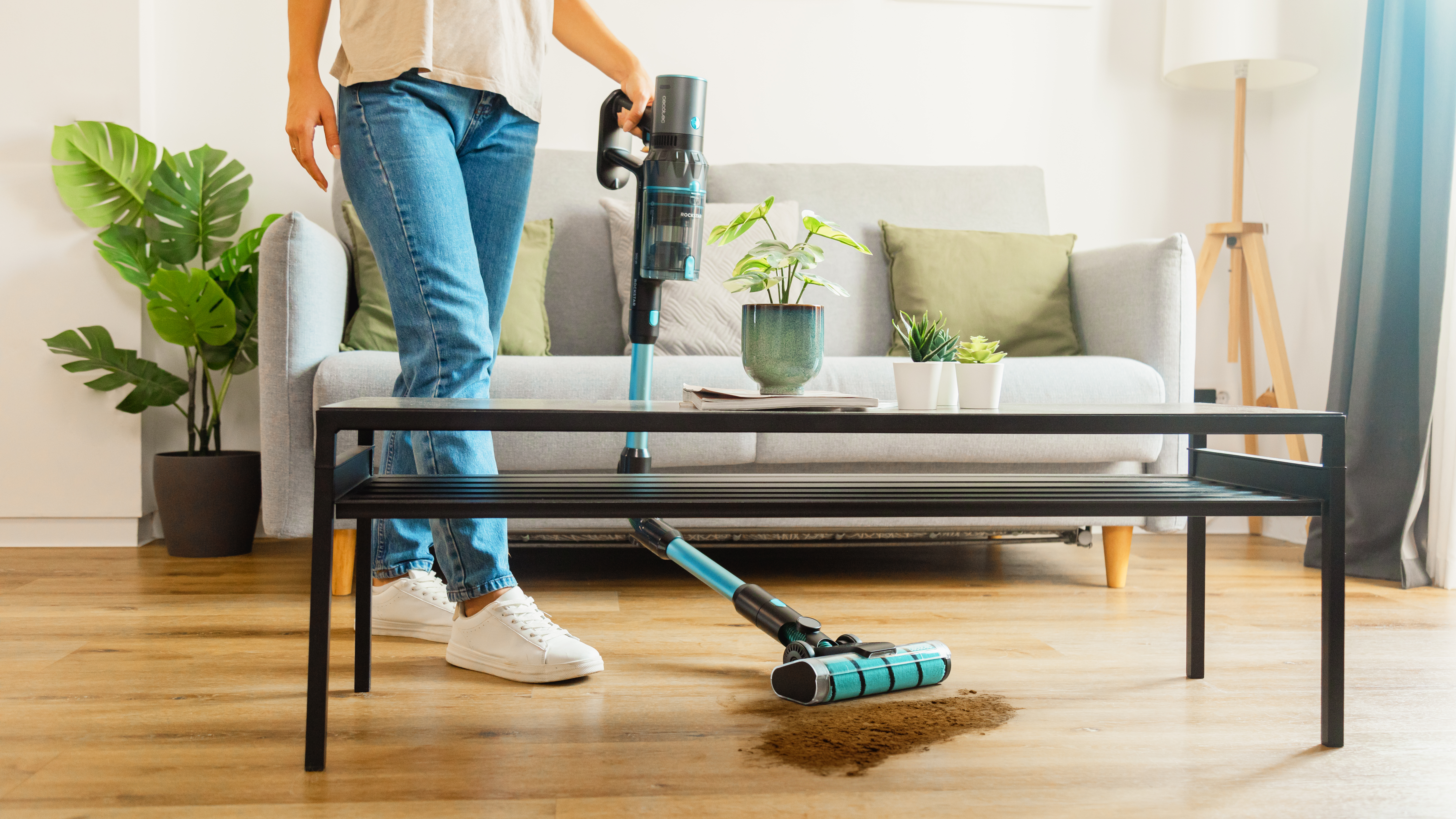 Una mujer aspira un piso de madera con una aspiradora inalámbrica. El suelo tiene un montón de suciedad y muebles detrás.