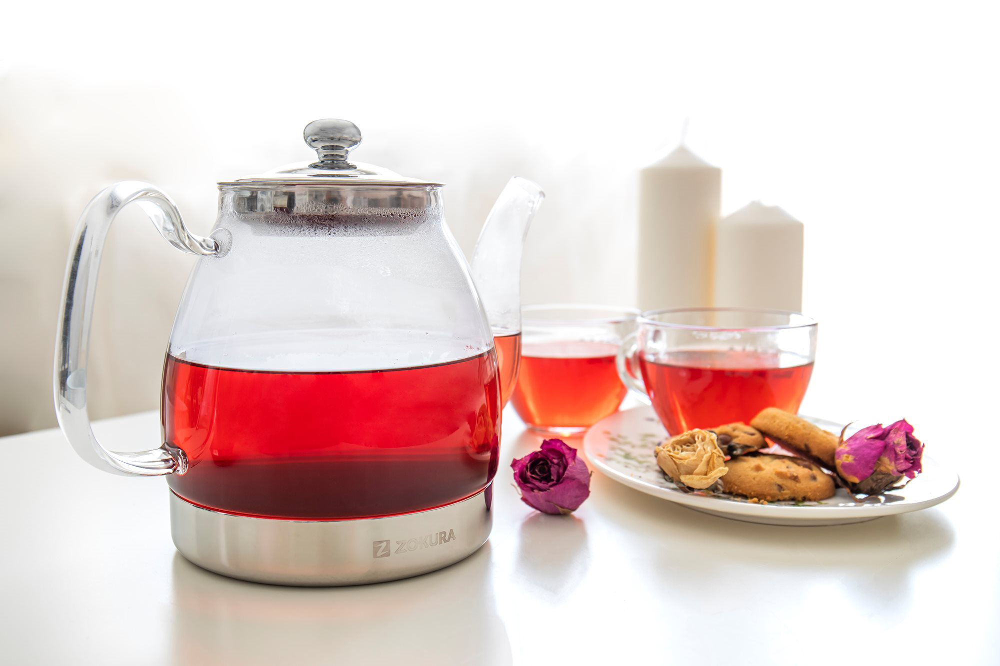 Egy üveg teáskanna piros folyadékkal, két csésze tea, sütemények és gyertyák egy fehér asztalon.