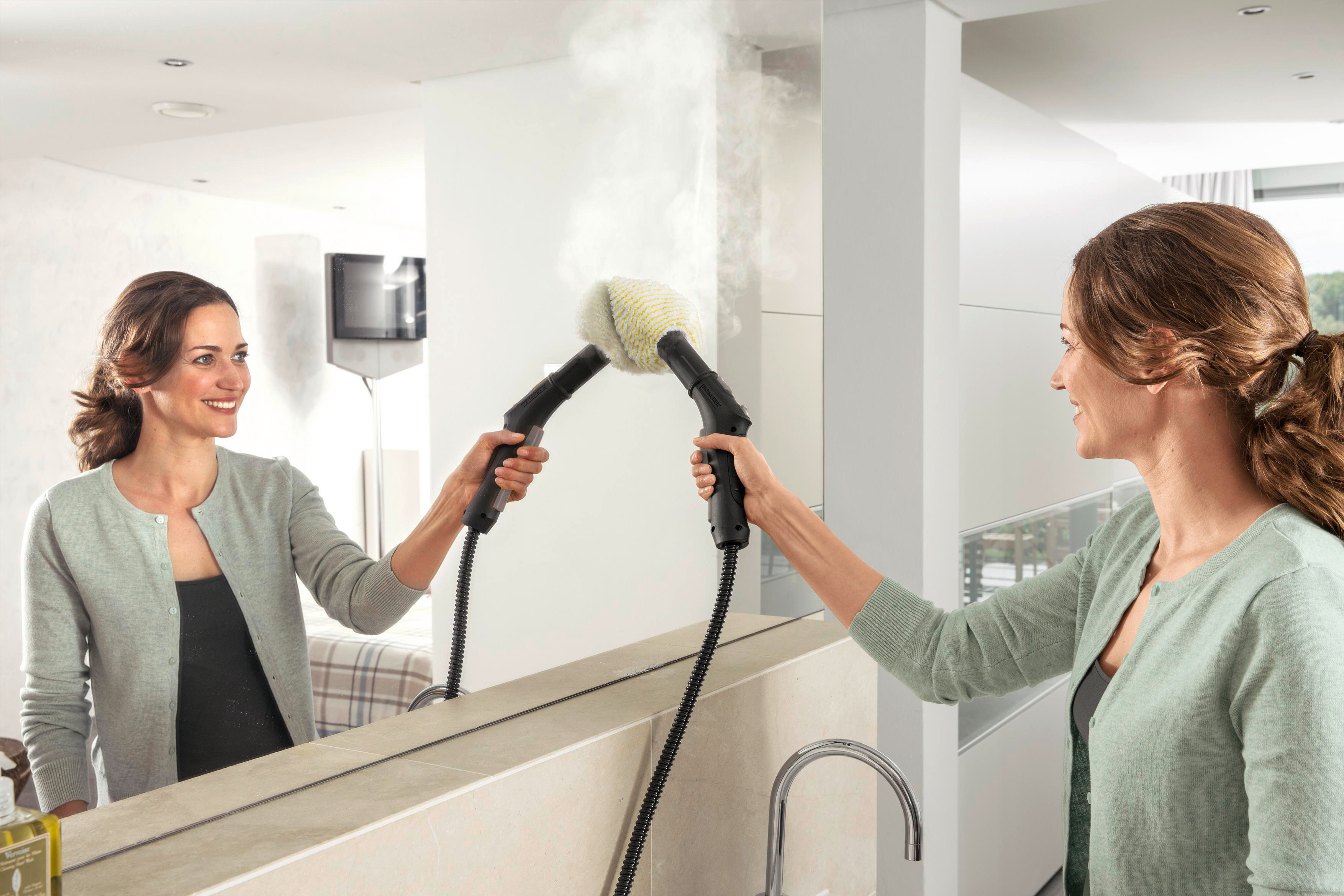 Zwei Frauen reinigen einen Spiegel mit einem Dampfreiniger, Spiegelung sichtbar, im Badezimmer.