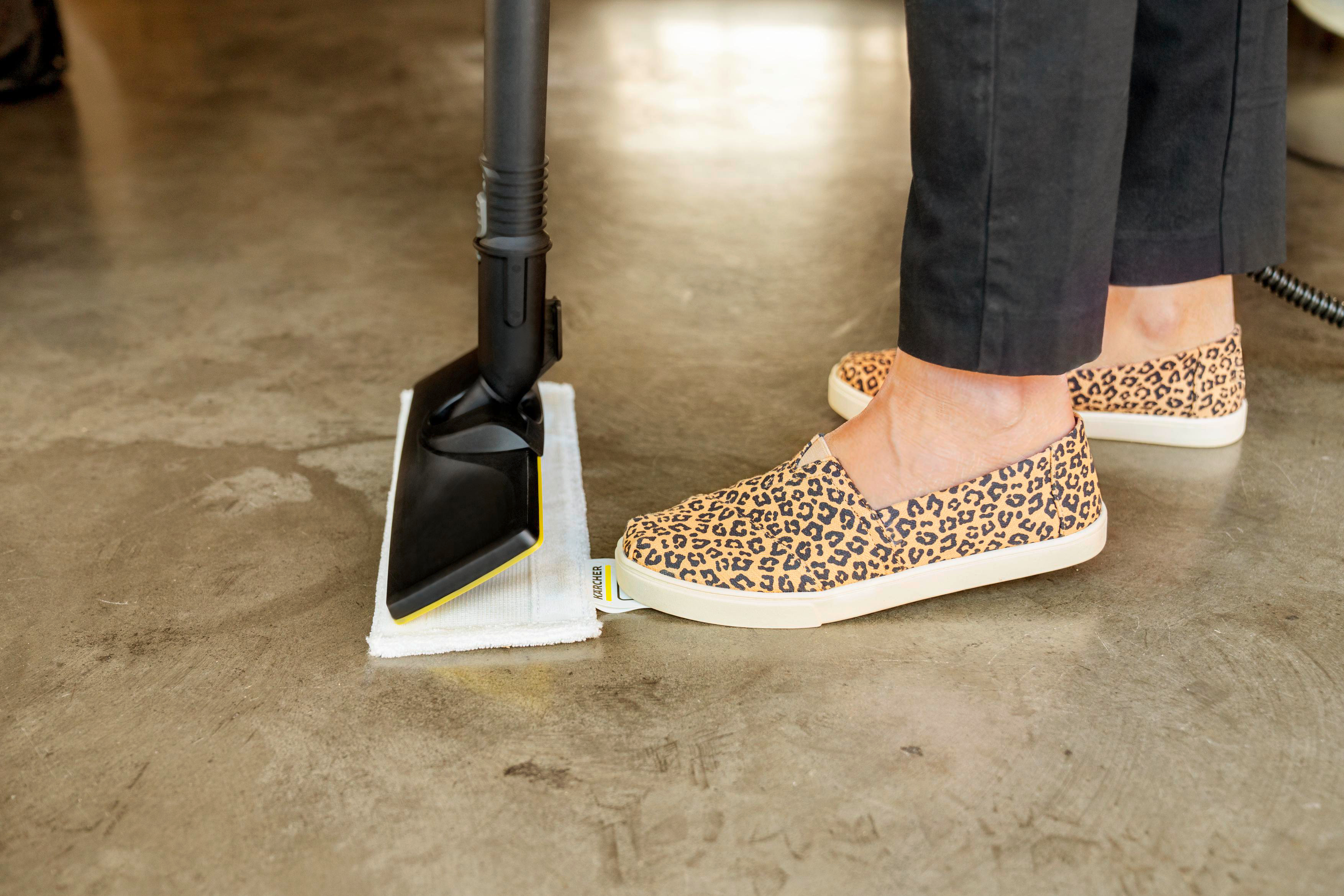 Personenfüße mit Leopardenmuster-Schuhen in der Nähe eines Dampfmopps auf Betonboden.