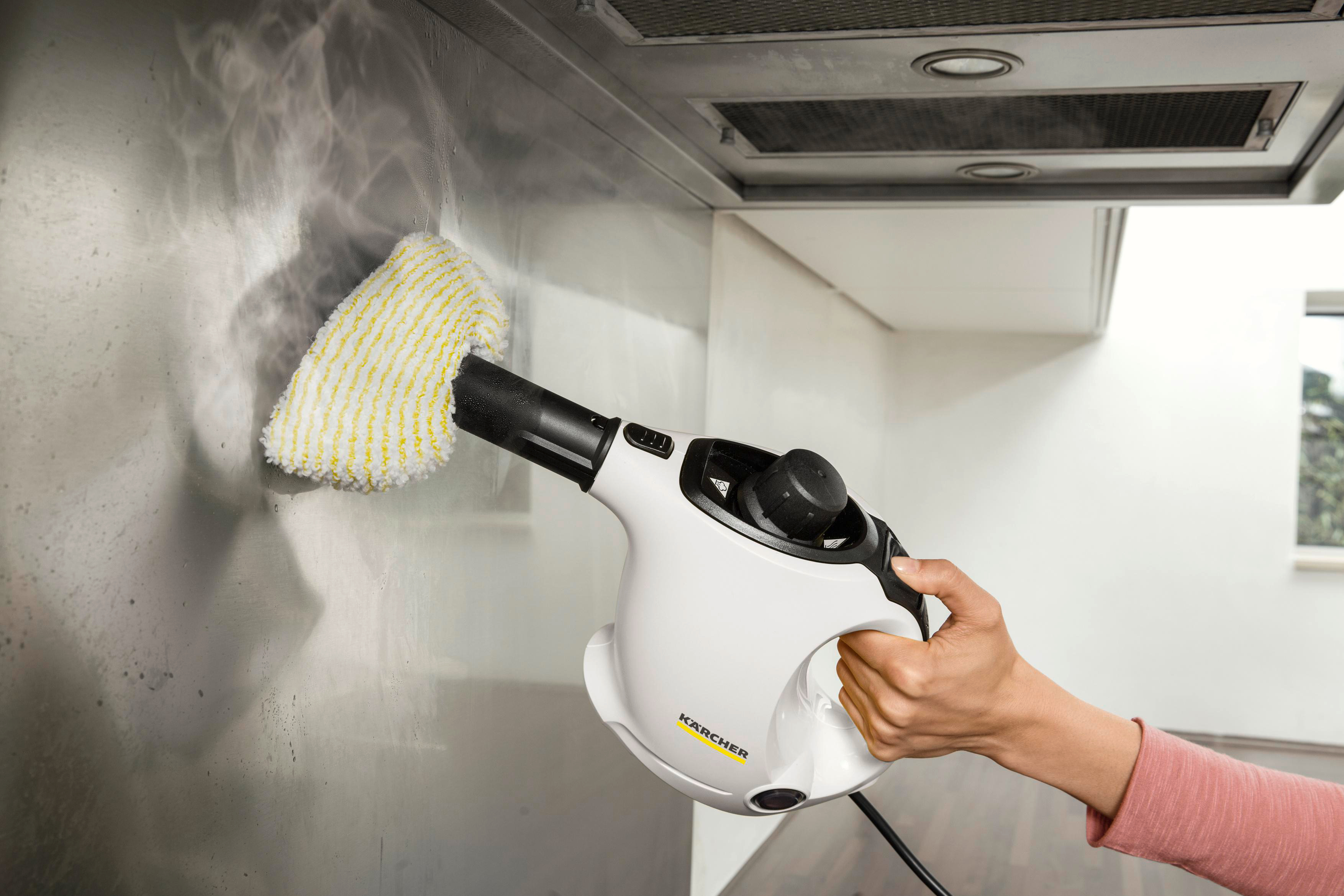 Eine Hand hält einen weißen Dampfreiniger gegen eine metallische Oberfläche, Dampf steigt auf.