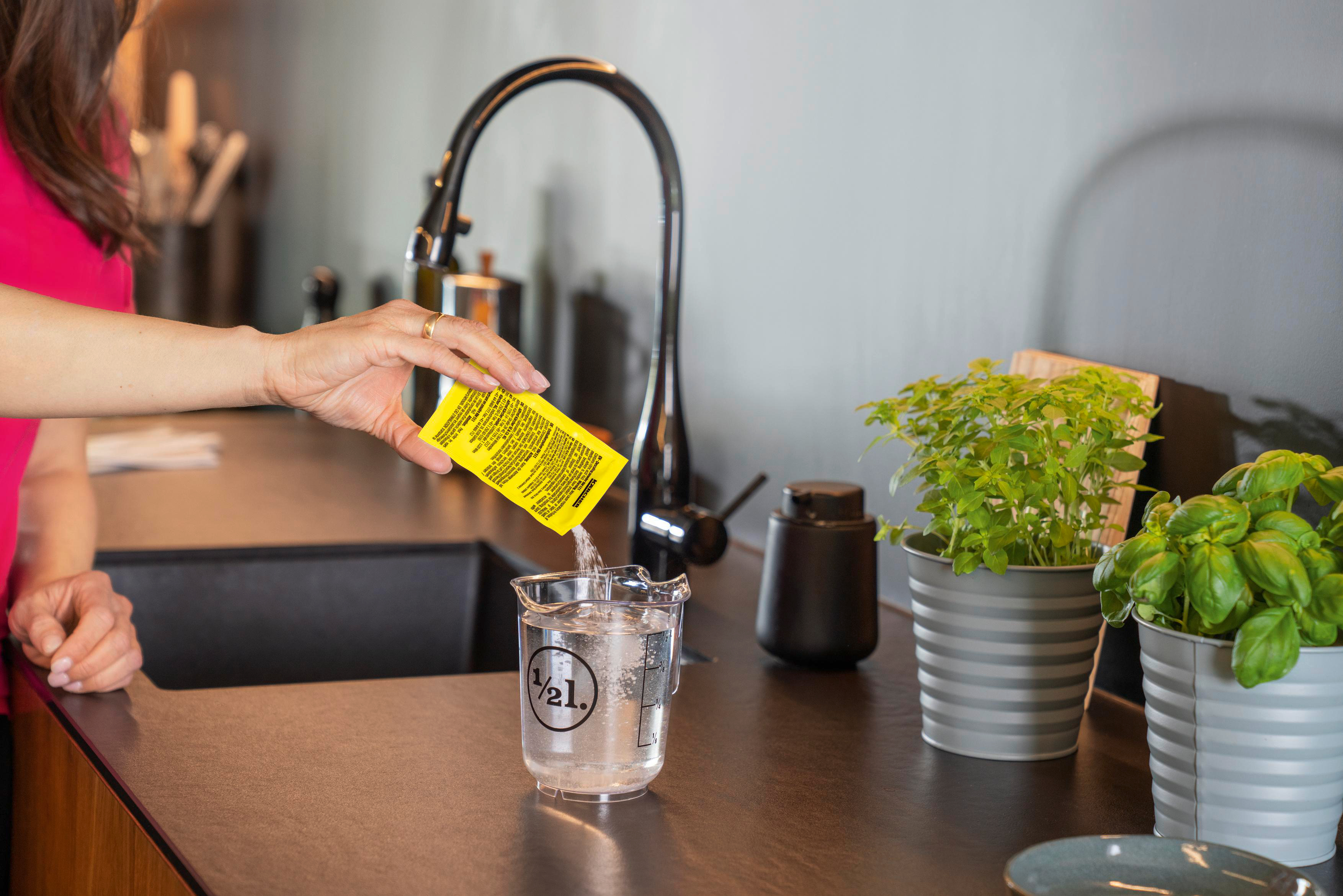 Hand, die ein Pulver aus einem gelben Päckchen in einen Becher Wasser auf einer Theke gießt.