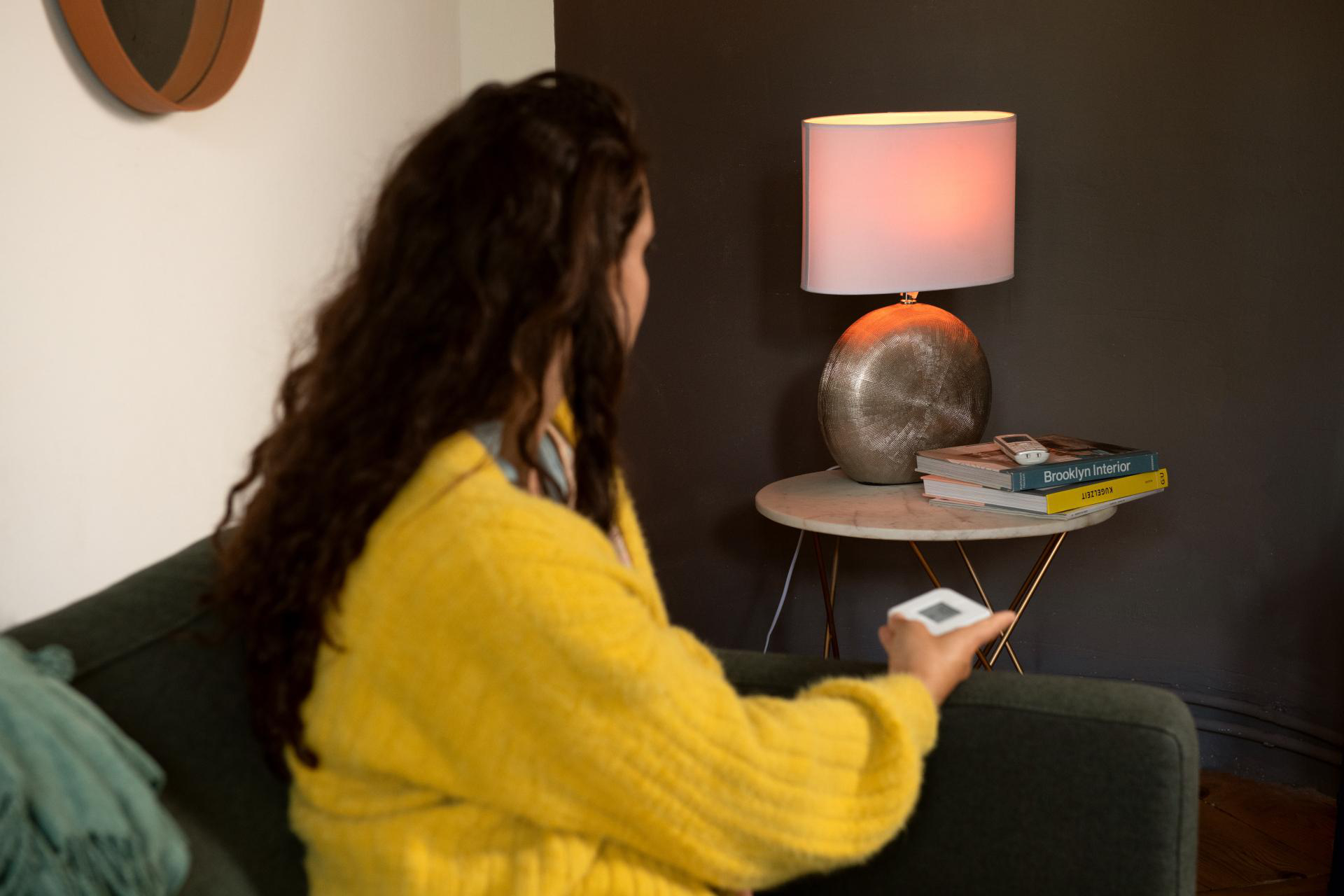 Frau mit gelber Jacke sitzt auf Sofa. Lampe auf Beistelltisch. Fernbedienung in der Hand.