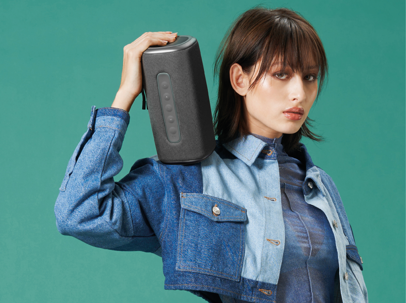 Femme tenant un haut-parleur gris foncé, veste en jean, fond turquoise.