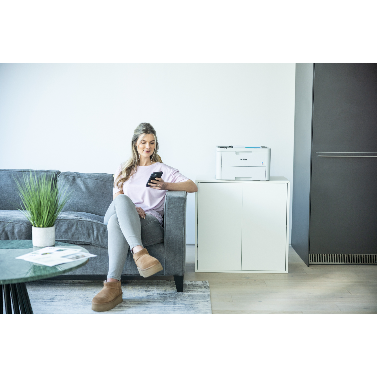 A woman sits on a gray sofa with a phone, near a white cabinet and a printer.