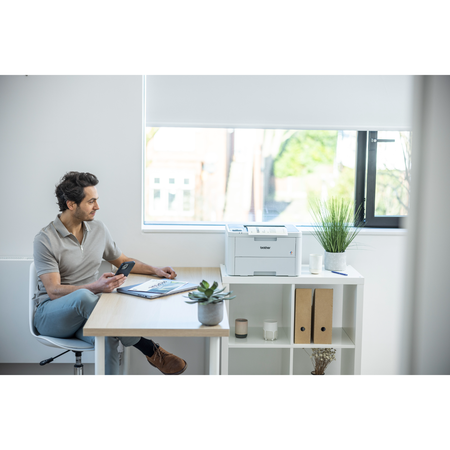 Man at desk with laptop and phone. A printer and a plant are also on the desk. White room.
