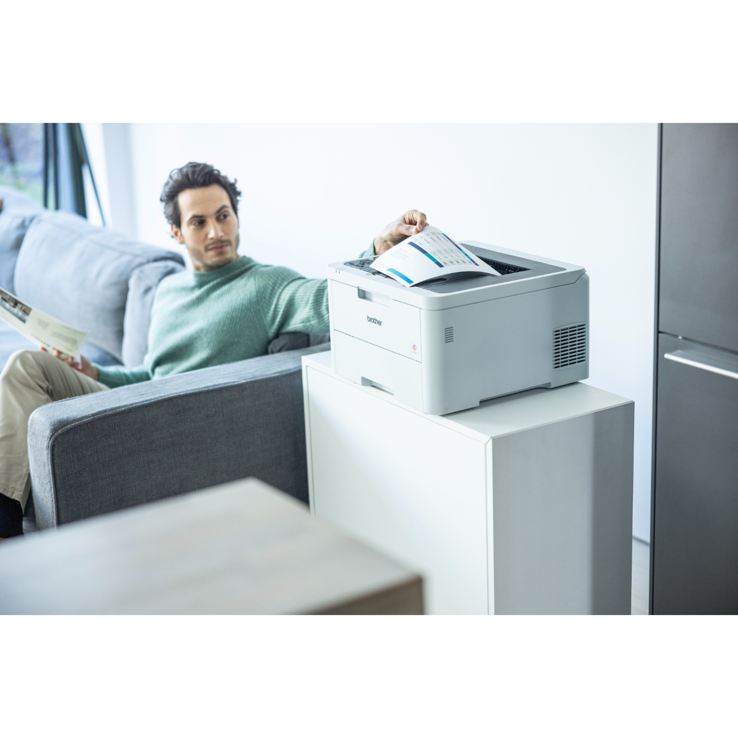 A man sits on a couch looking at a magazine. A printer is on a white cabinet next to him.