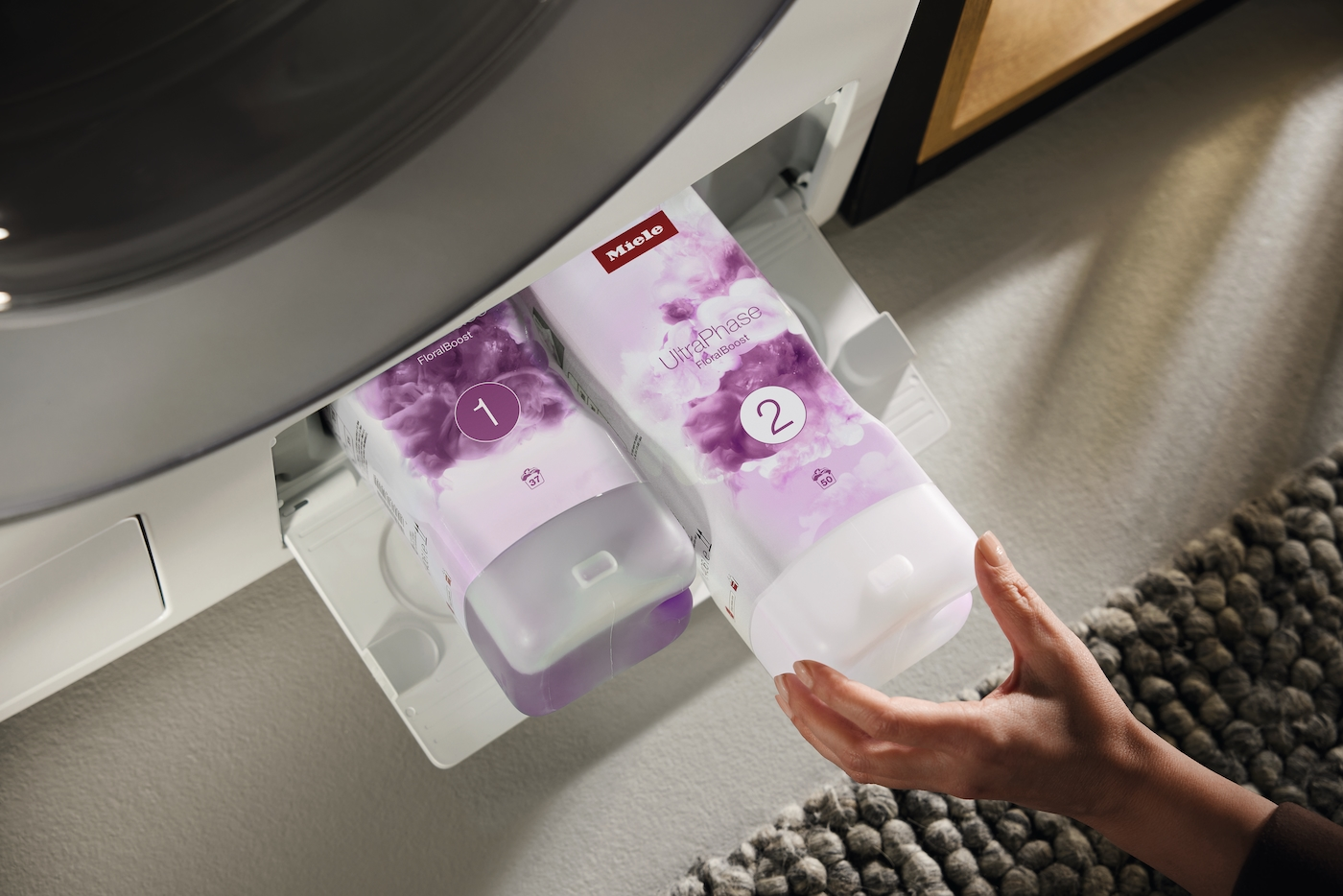 Zwei Miele Waschmittelkartuschen in einer Schublade. Eine Hand hält Kartusche 2.