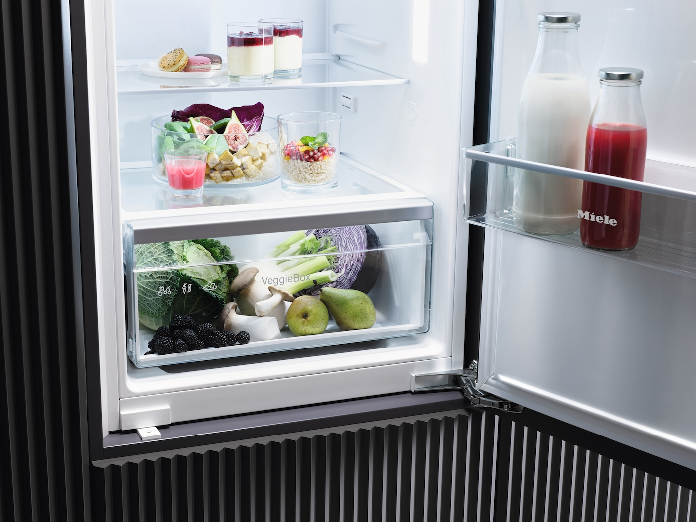 Geöffneter Kühlschrank, zeigt Gemüse, Obst und Getränke in Glasbehältern. Miele Logo.