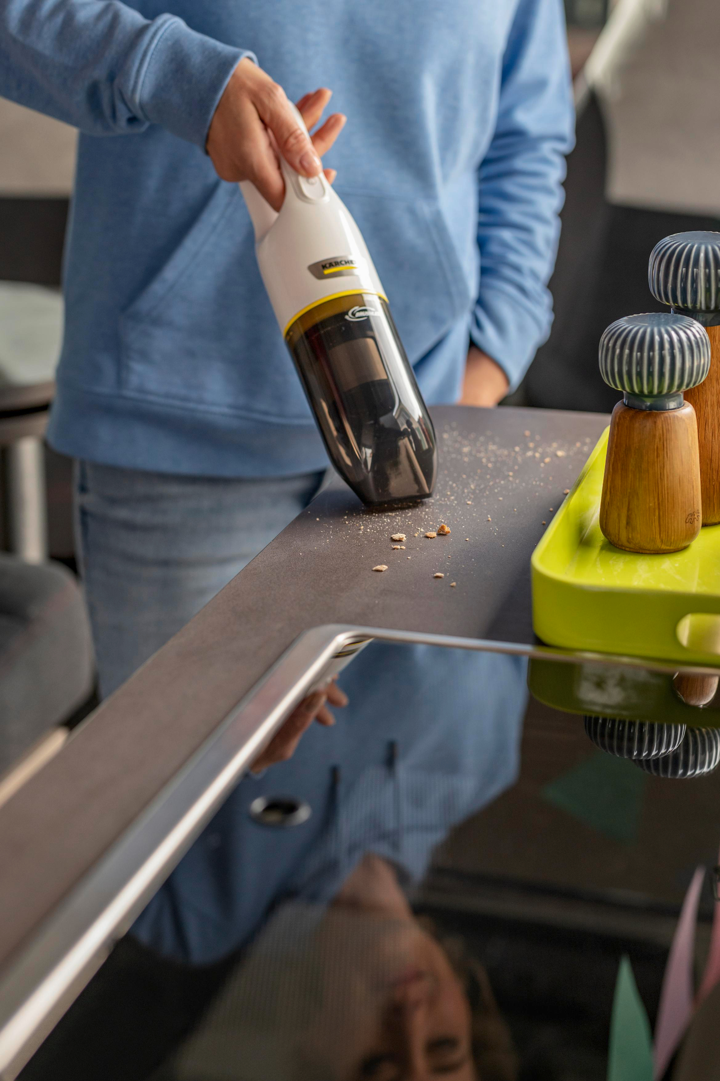 Eine Hand staubsaugt einen Tisch, Krümel und zwei Salzstreuer sind sichtbar.