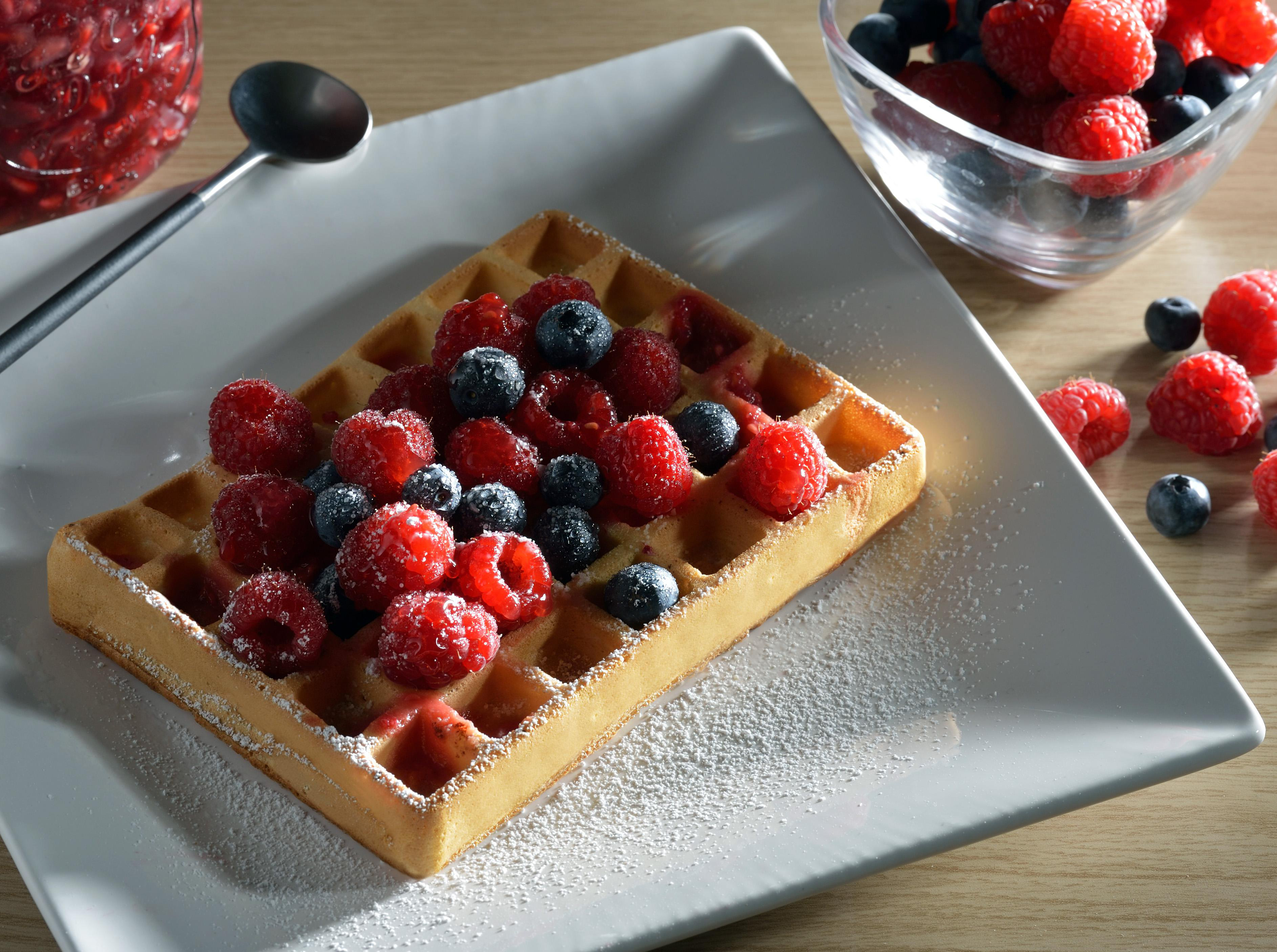Waffel mit Beeren und Puderzucker auf einem weißen Teller, mit Löffel und Marmelade daneben.