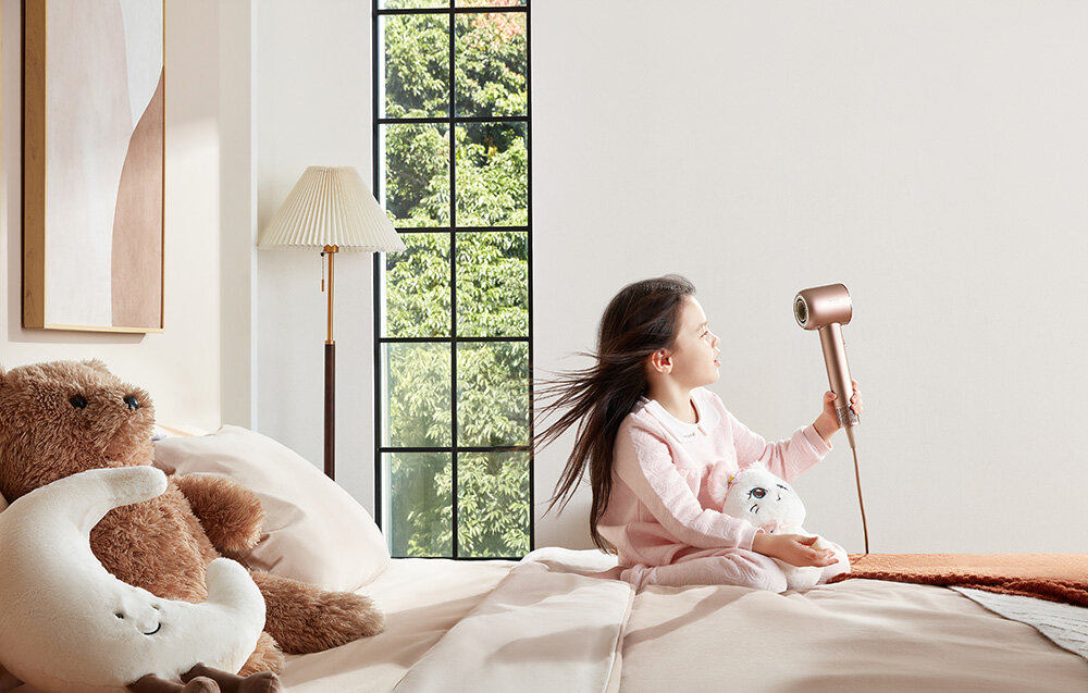 Dziewczynka trzyma suszarkę do włosów przy oknie, miś i księżyc na łóżku.