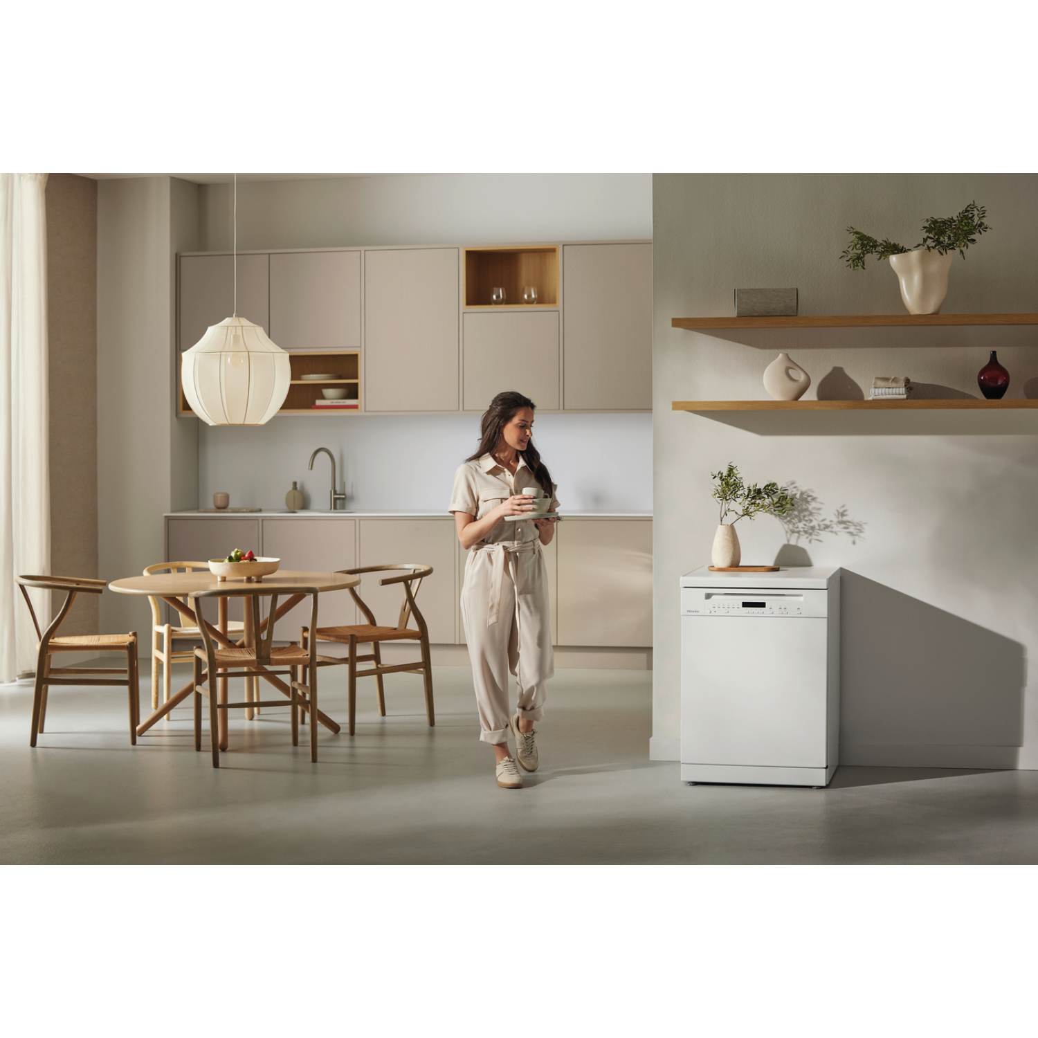 Een vrouw in een keuken met een kopje, met een vaatwasser en eettafel zichtbaar, in een lichte kamer.