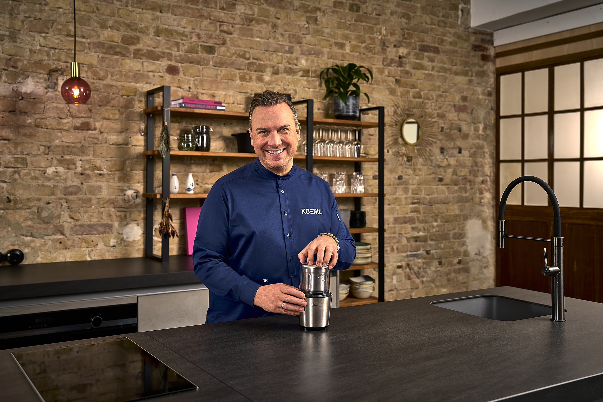 Ein Mann in blauem Hemd, lächelnd, bereitet Kaffee mit einem silbernen Kaffeezubereiter zu.
