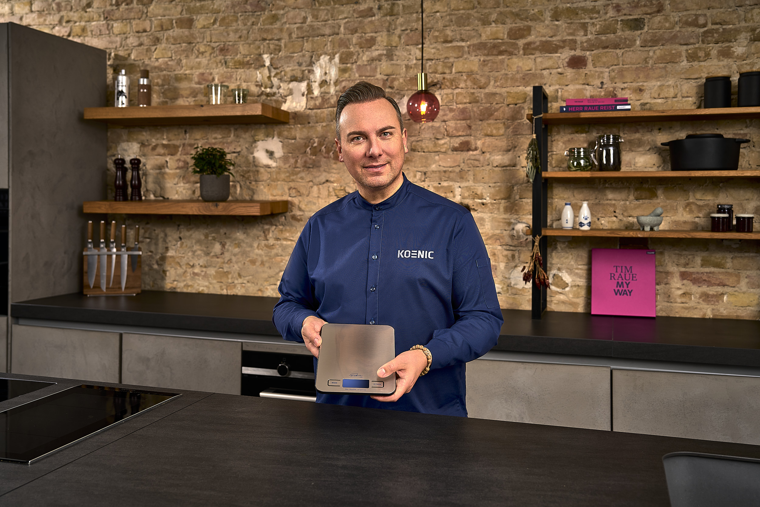 Ein Mann in blauem Hemd hält eine Waage in einer modernen Küche. Ziegelwand im Hintergrund.