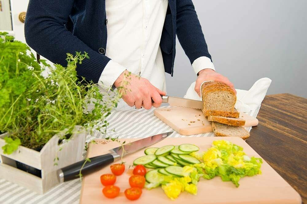 Osoba kroi chleb obok warzyw. Tło: deska do krojenia, kuchnia i rośliny.