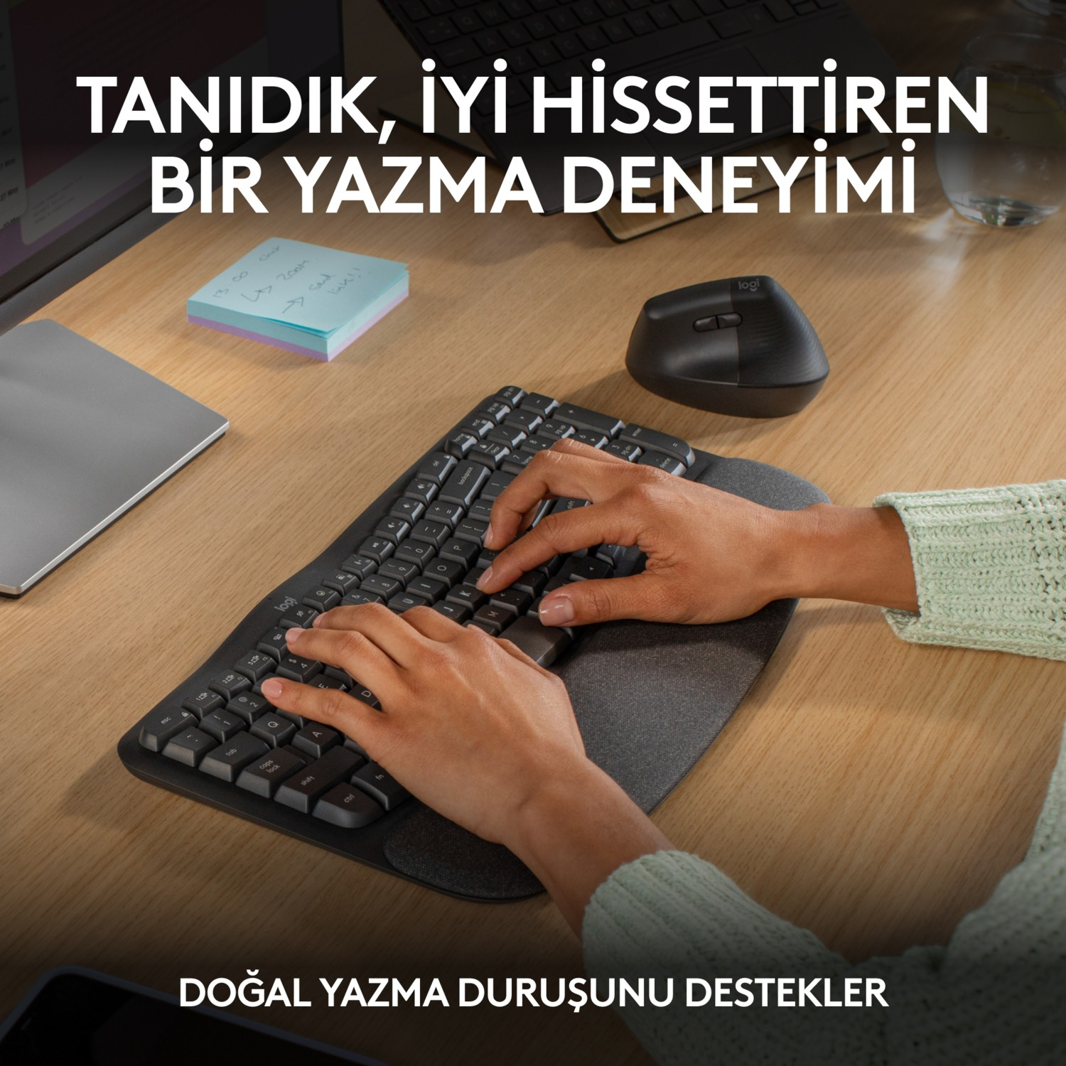 Eller, bilek desteği olan siyah bir klavyede yazıyor. Siyah bir fare ve yapışkan notlar görünür.