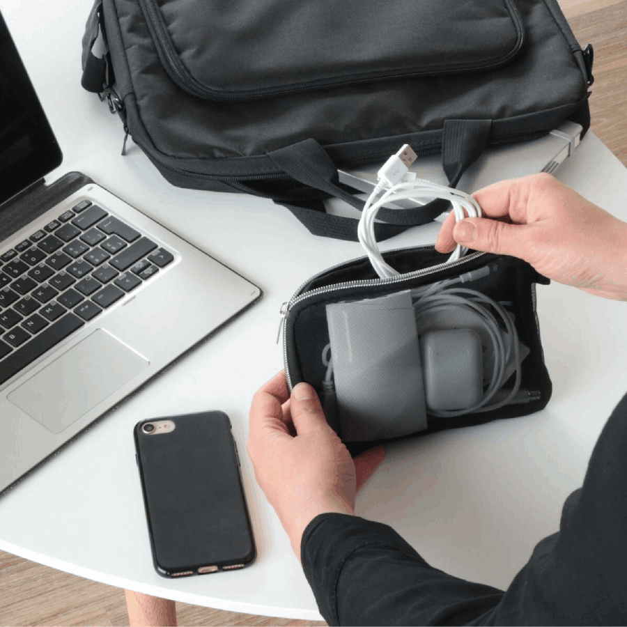 Manos sosteniendo una pequeña funda con cables, portátil, teléfono y bolso en mesa blanca.