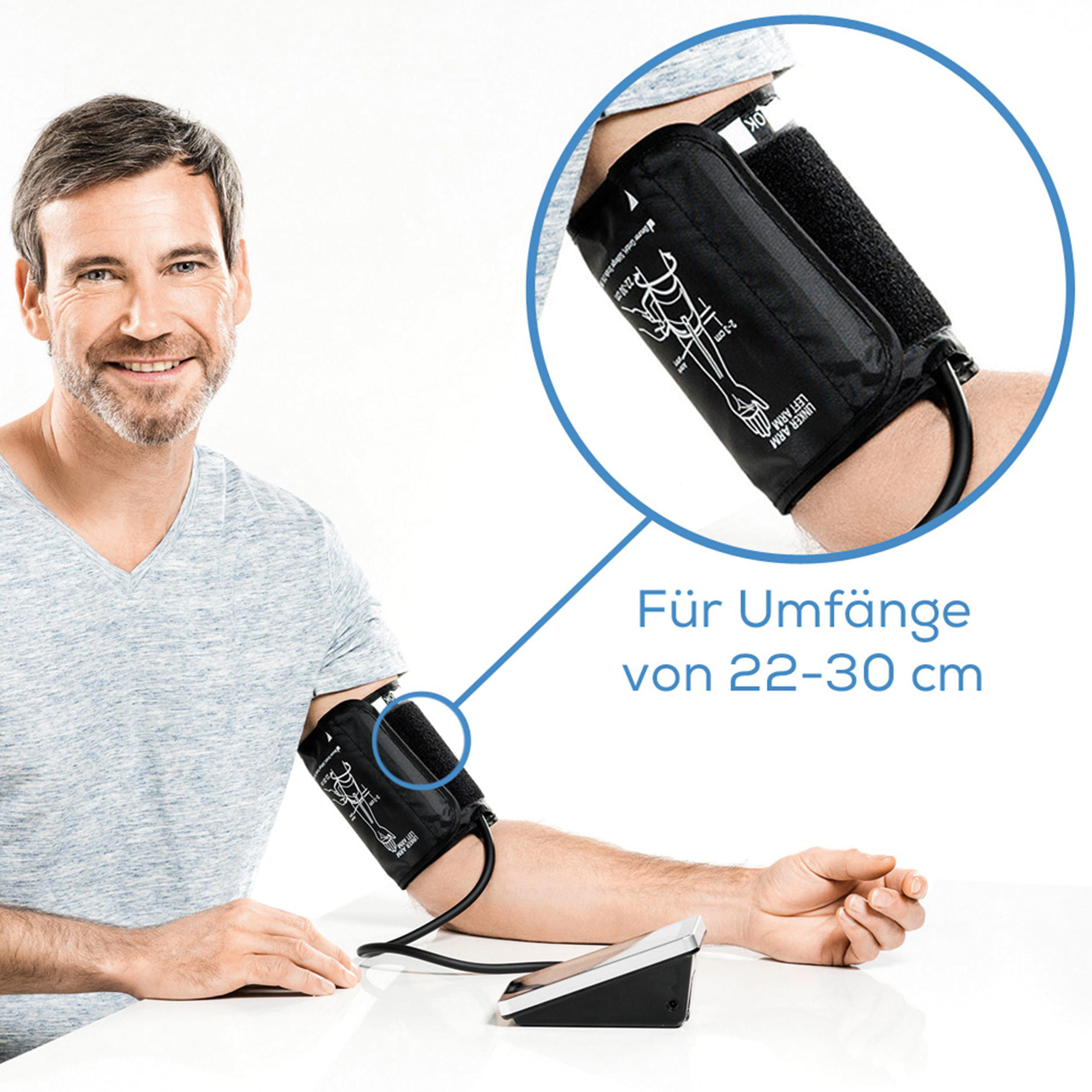 Mann, der seinen Blutdruck misst, mit einer schwarzen Manschette am Arm, lächelt in die Kamera.