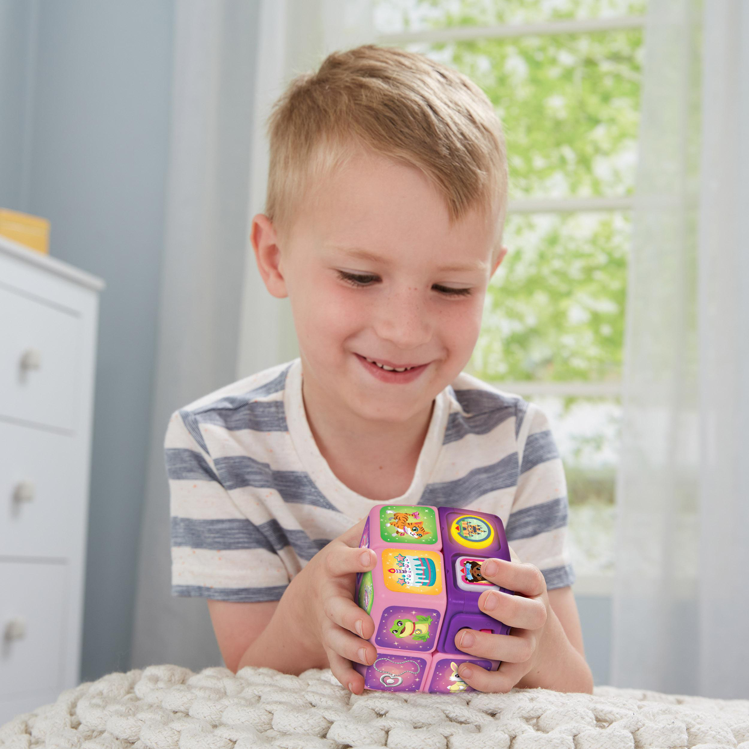 Ein kleiner Junge hält ein buntes Würfelspielzeug und lächelt. Er ist in einem Raum mit einer weißen Kommode.
