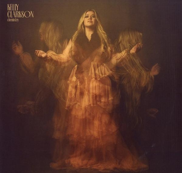 Albumcover von Kelly Clarkson, in einem orangefarbenen Kleid, mehrere verschwommene Figuren, auf dunklem Hintergrund.