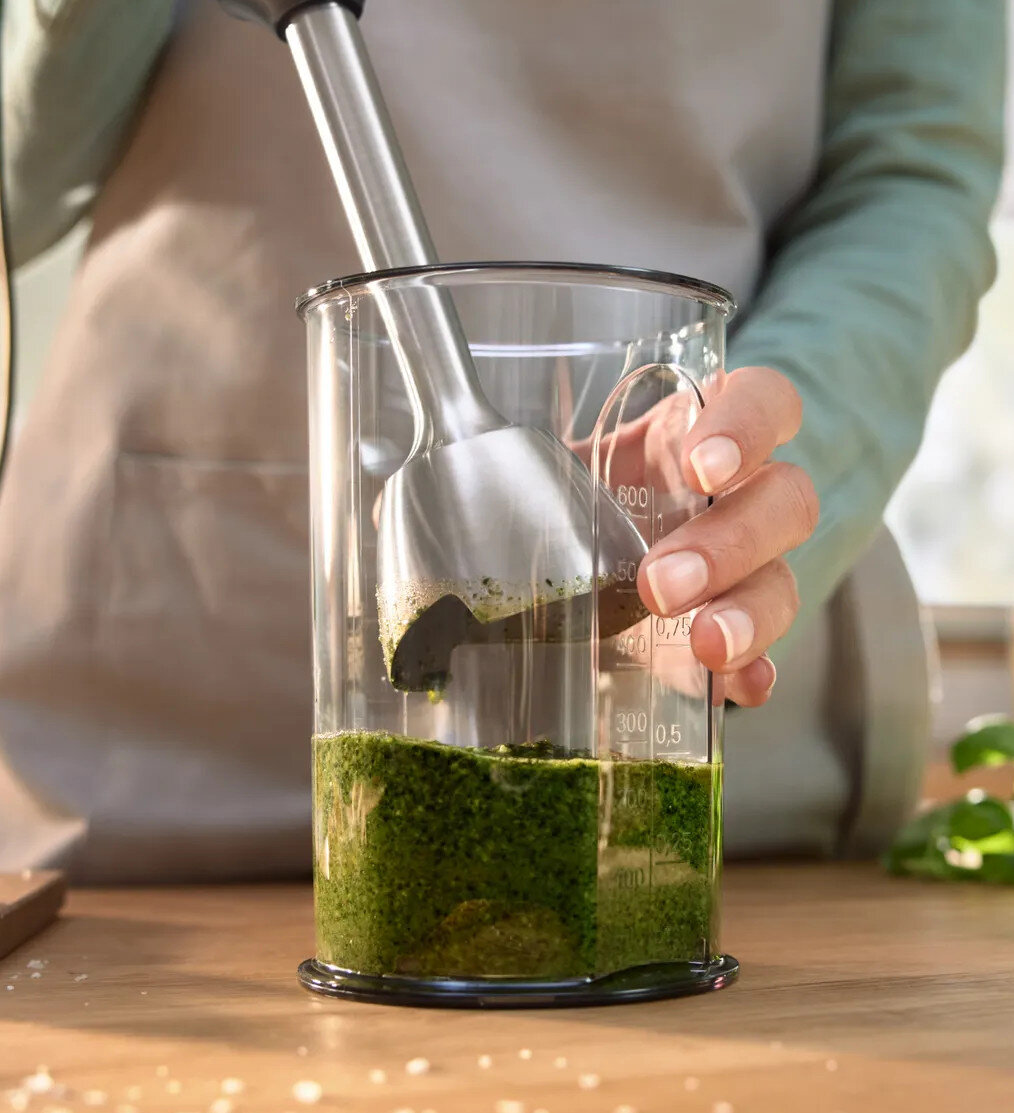 Ręka trzyma blender ręczny w szklanym pojemniku z zielonym sosem. Jasne tło.