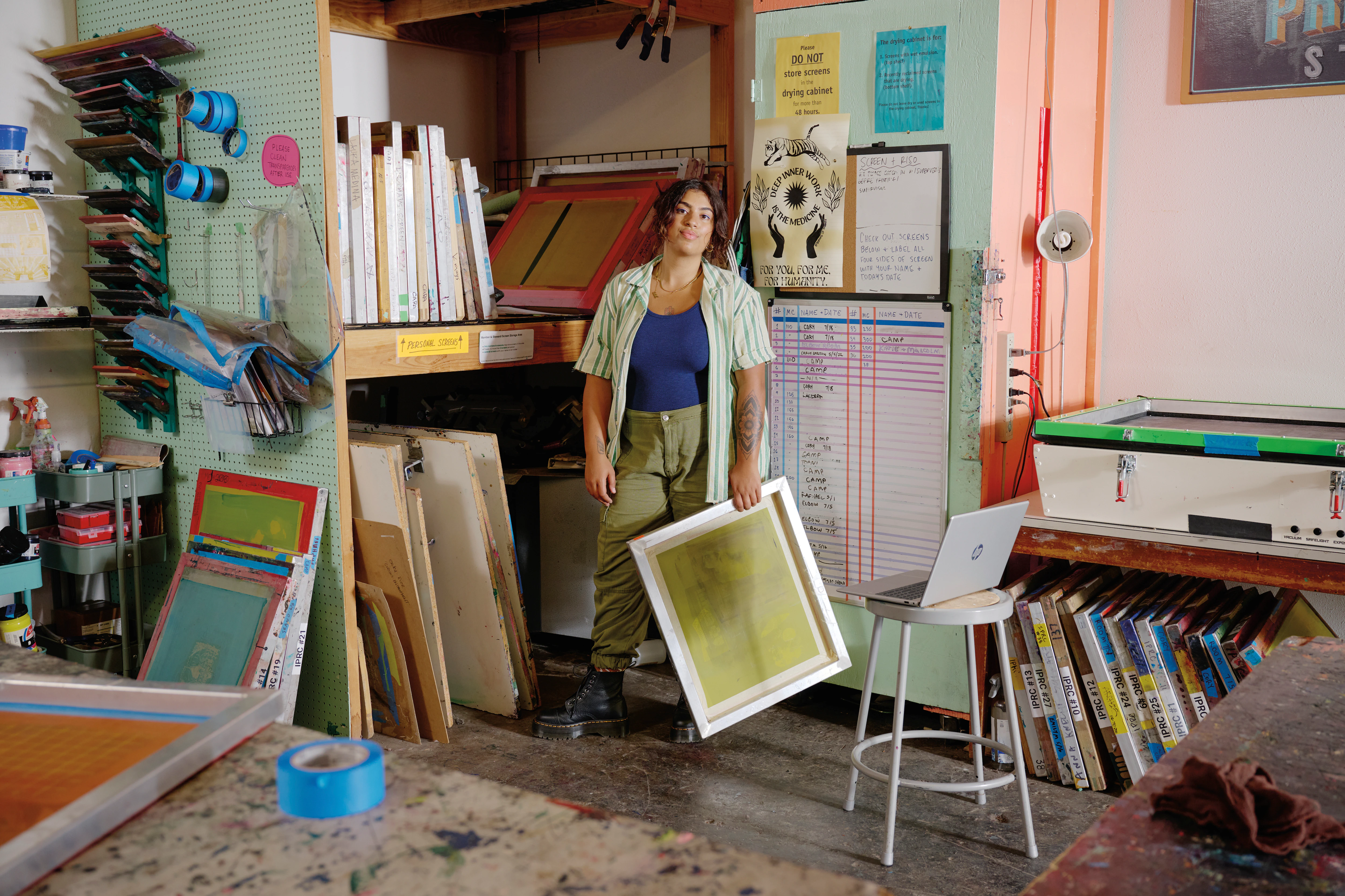 Mujer en un estudio sosteniendo un marco de serigrafía, con laptop y suministros.