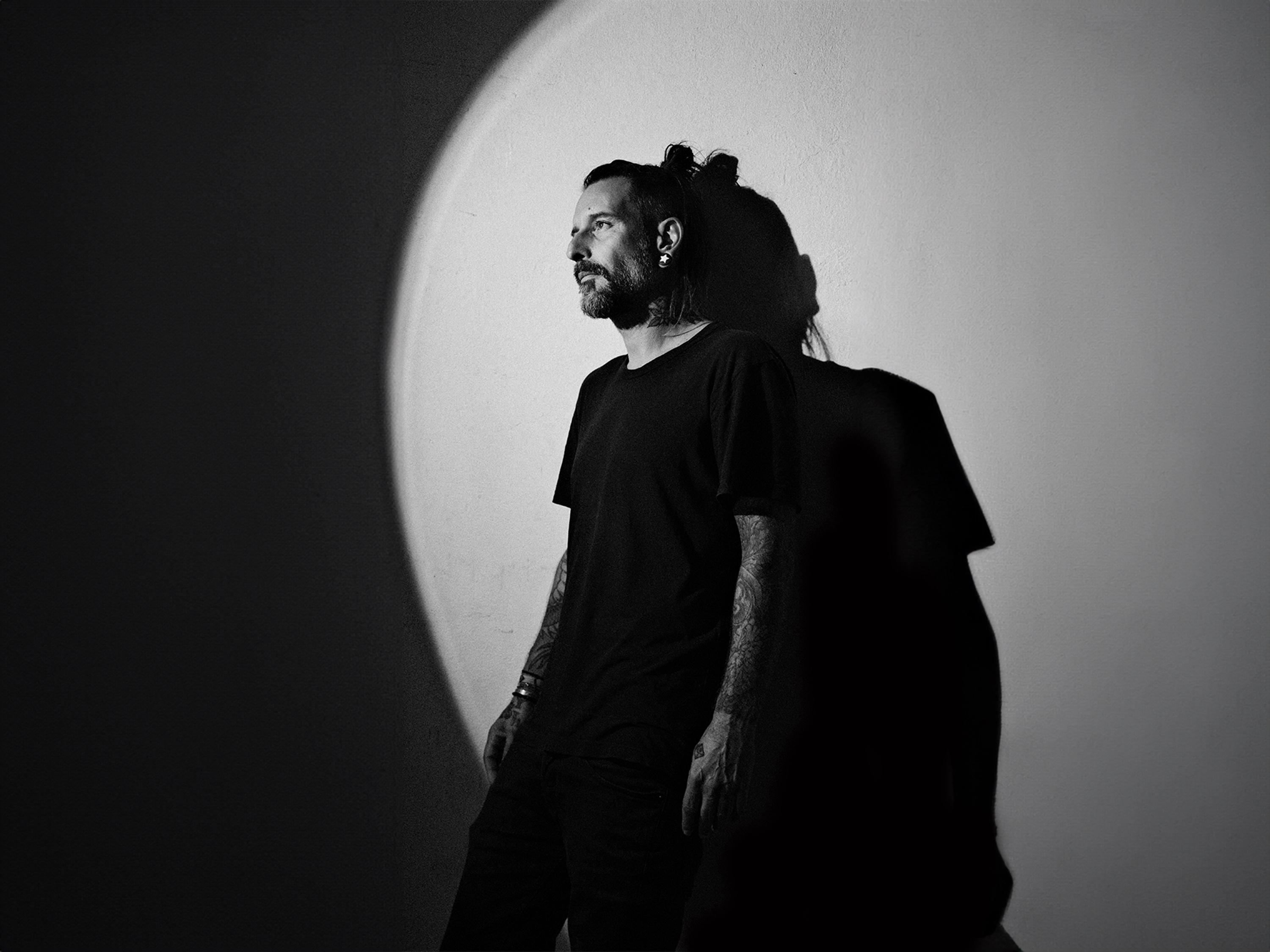 Mann in schwarzem T-Shirt und Hose im Scheinwerferlicht. Schatten an der Wand hinter ihm.