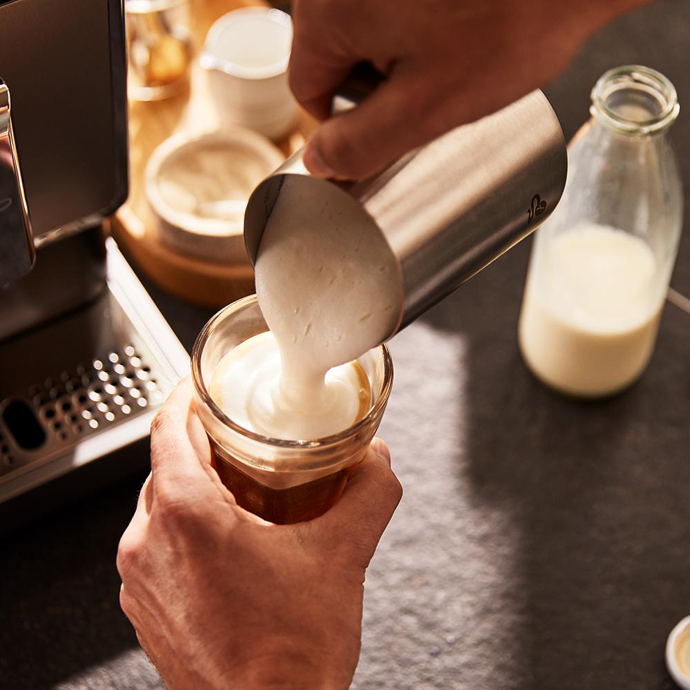 Milchschaum in ein Glas Kaffee gießen. Andere Zutaten stehen auf der Theke.