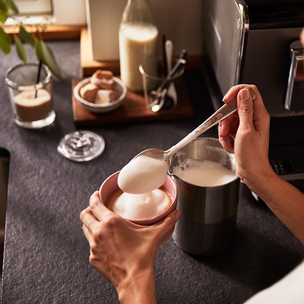 Eine Person schöpft Schaum in eine rosa Kaffeetasse, mit verschiedenen Kaffeezubereitungswerkzeugen.