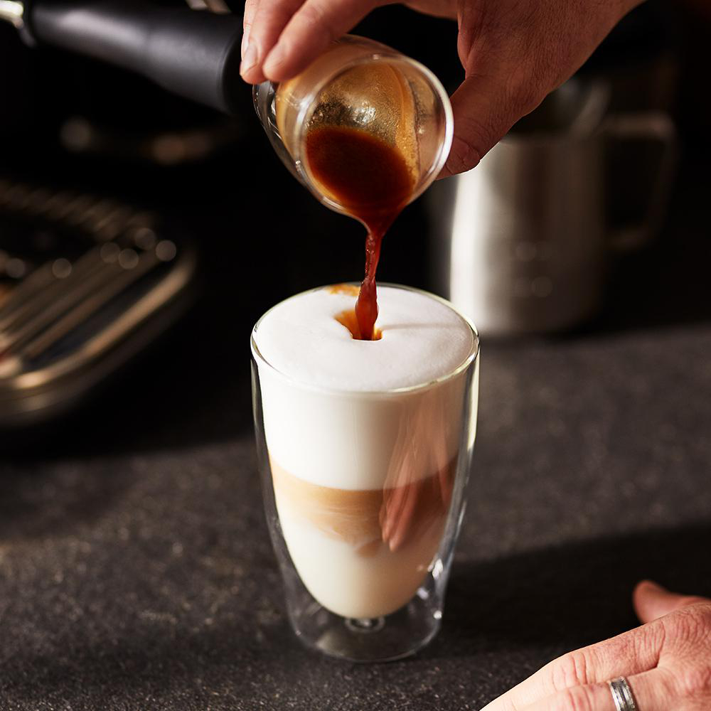 Eine Hand gießt Espresso in ein Glas Milch und erzeugt so einen geschichteten Latte.