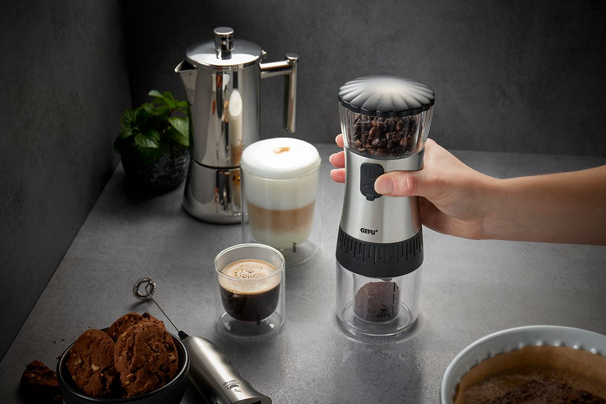Eine Frau mahlt Kaffeebohnen mit einer Handmühle, Kaffee und Gebäck im Bild.
