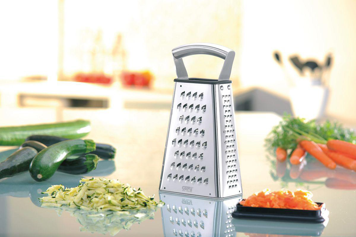 Eine Reibe mit geriebenen Zucchini und Karotten, mit Gemüse im Hintergrund.