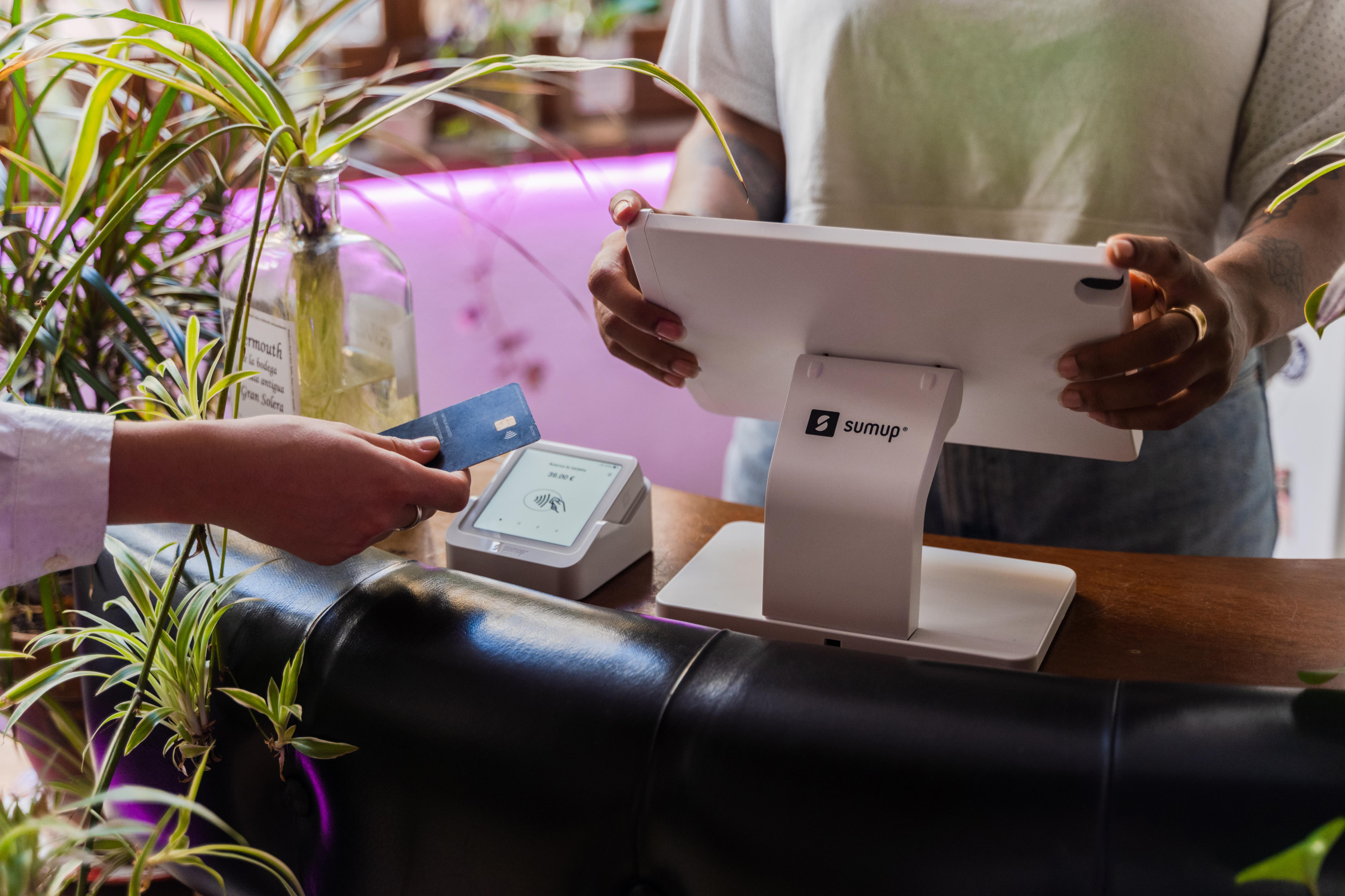 Eine Person, die mit Karte an einem POS-Terminal bezahlt, mit einer Pflanze im Hintergrund.