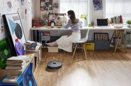 Kobieta siedzi przy biurku, robot odkurzający na podłodze. Książki, papiery i dekoracje. Białe tło.