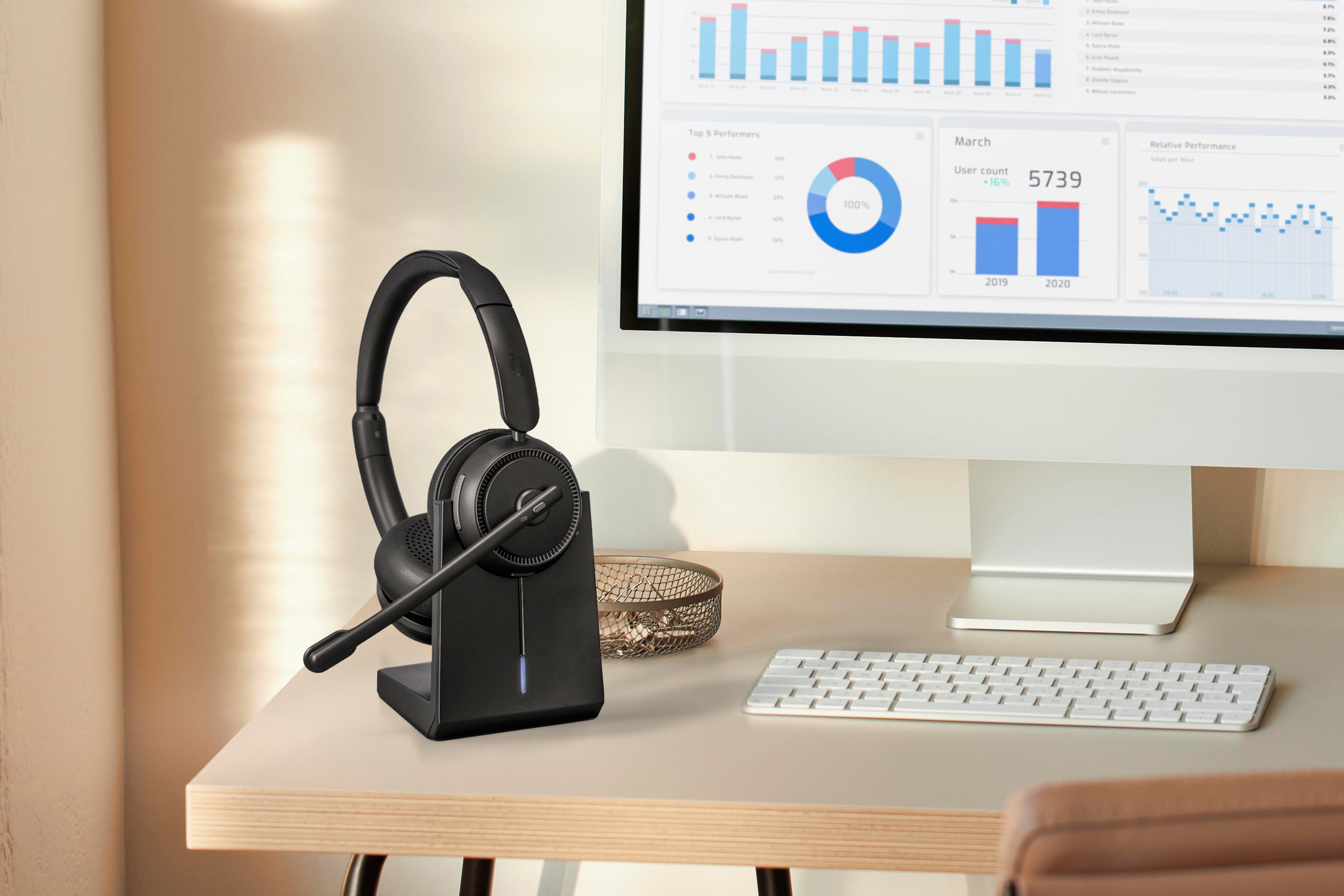 Ein schwarzes Headset auf einem Ladeständer steht auf einem Schreibtisch vor einem Monitor mit Diagrammen.