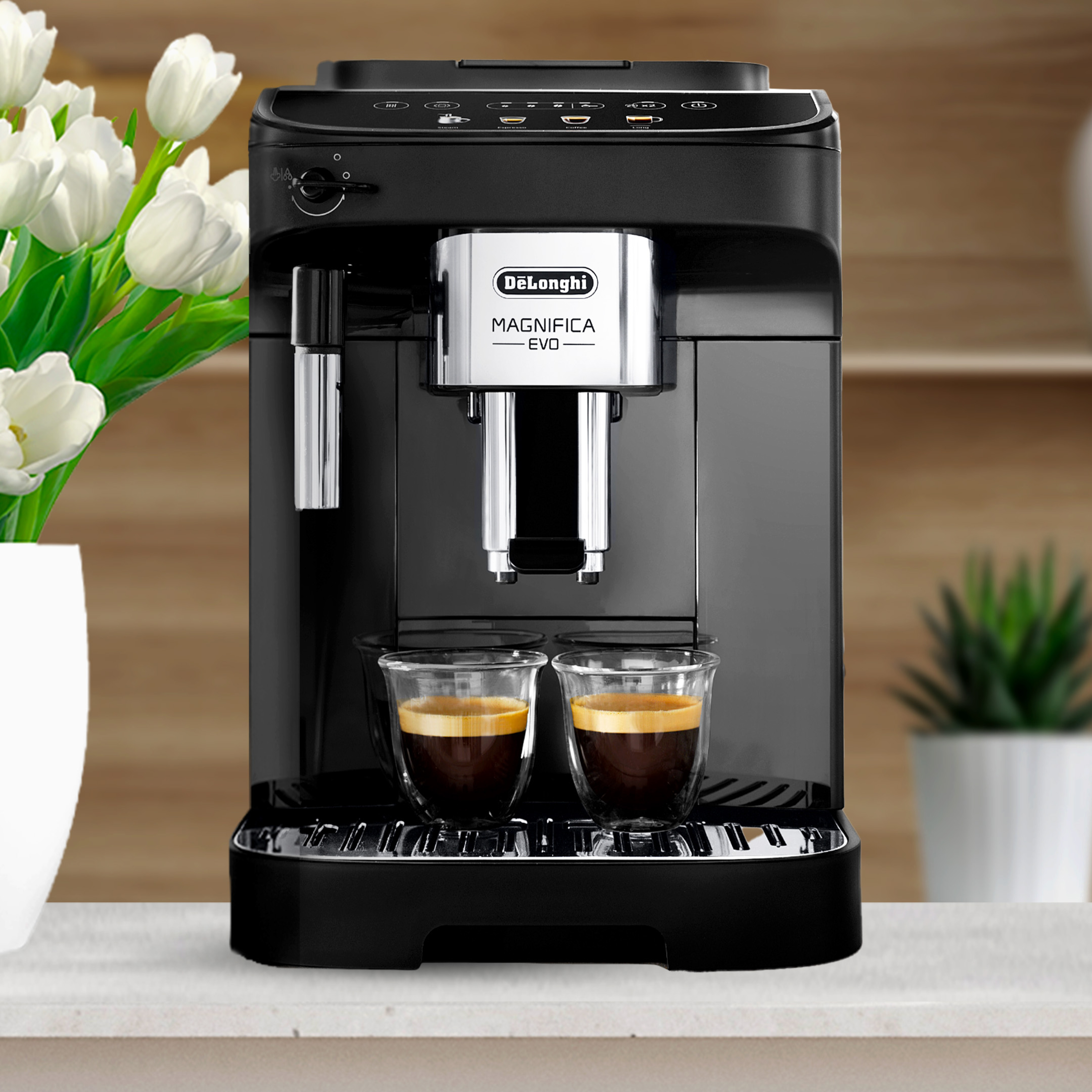 Una cafetera De'Longhi negra con dos tazas de café de vidrio y un jarrón de tulipanes blancos.