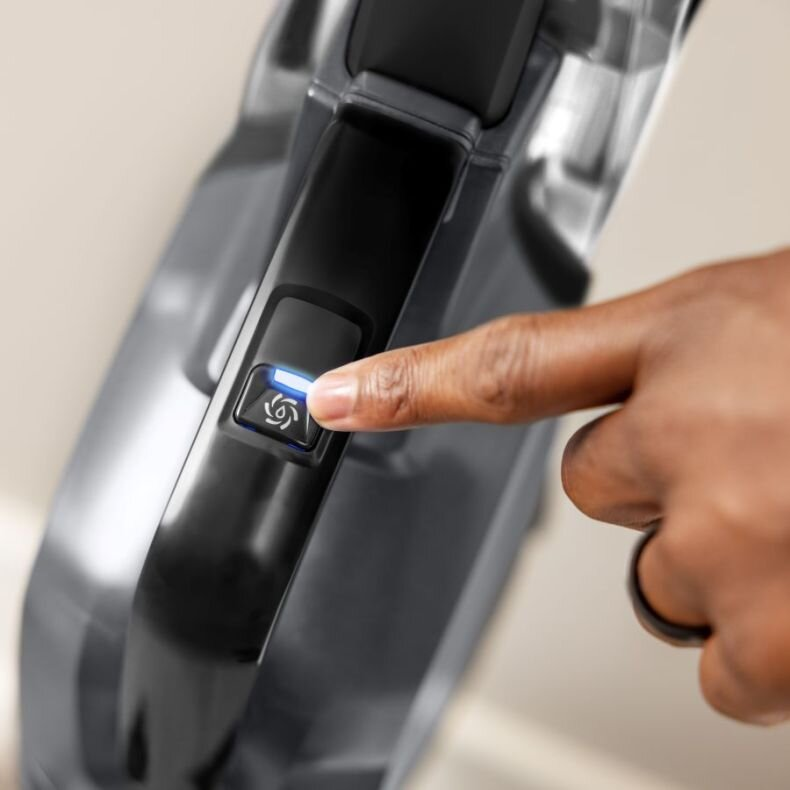 Palec naciska przycisk na czarnym odkurzaczu. Niebieskie światło wskazuje włączenie.