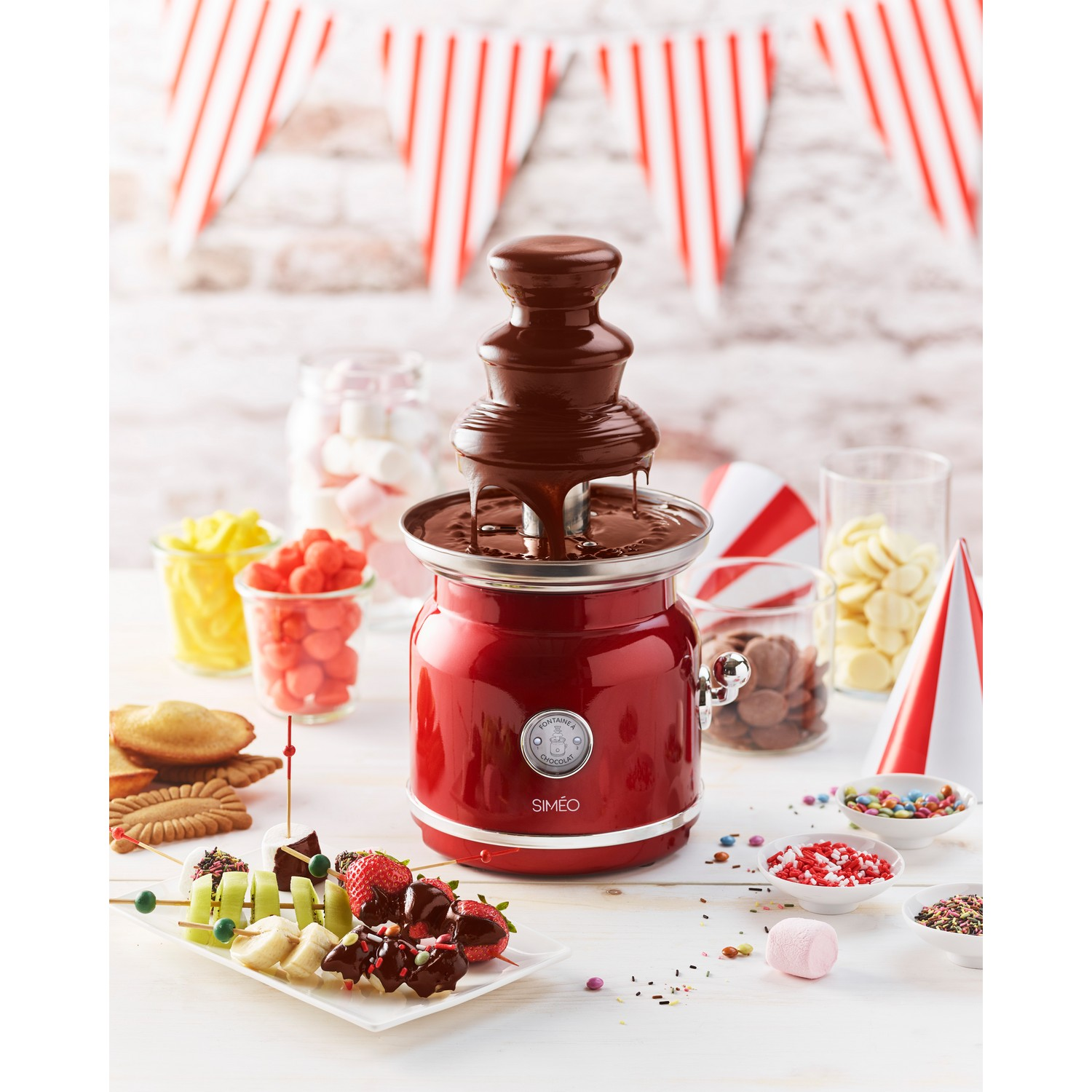 Chocoladefontein met een rode basis, omringd door lekkernijen, op een wit oppervlak.