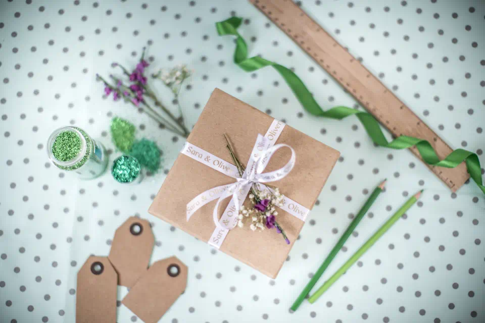 Boîte cadeau avec ruban, étiquettes, crayons et paillettes sur fond pointillé.
