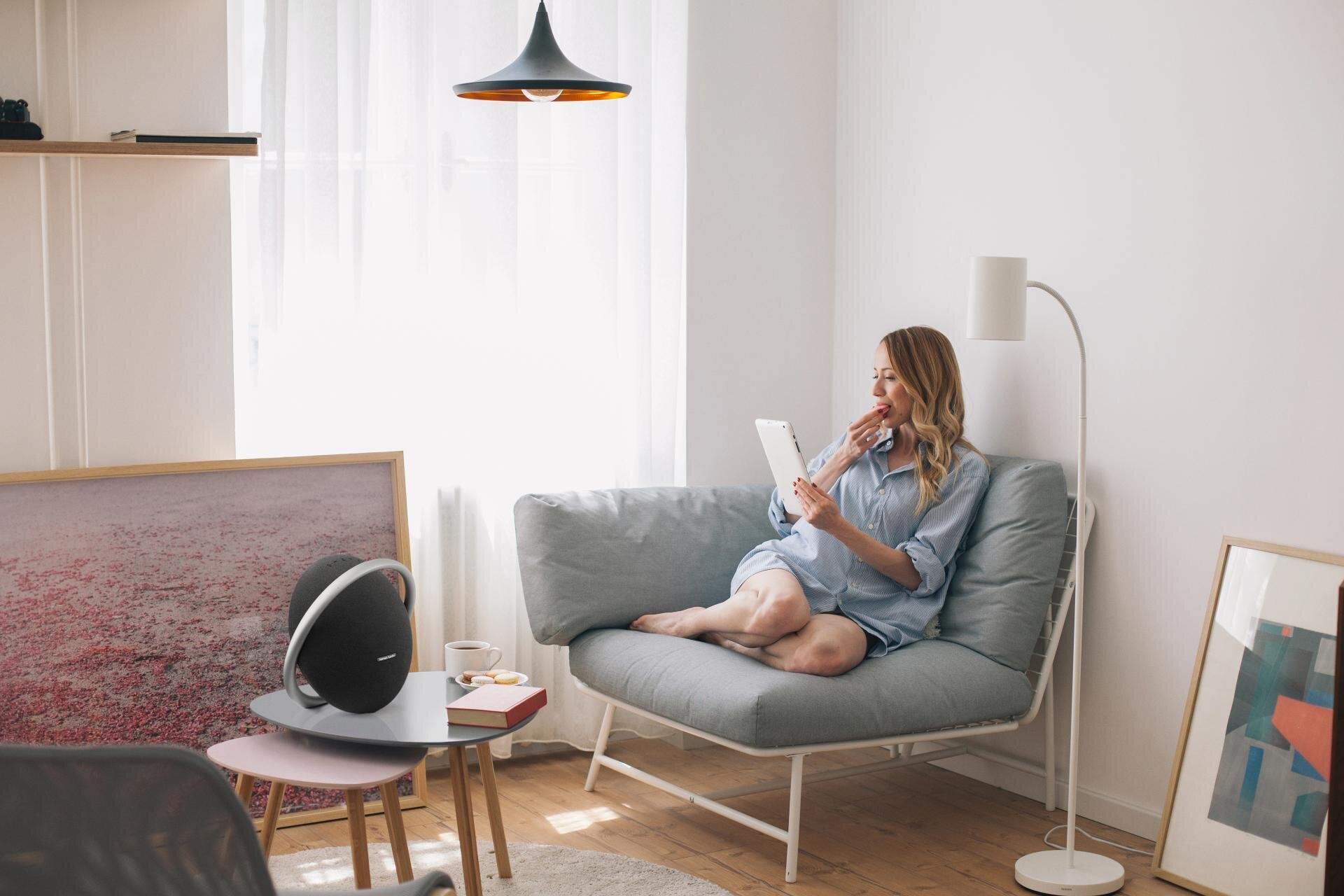 Kobieta z tabletem w pokoju, głośnik na stole. Sofa, okno, lampa i sztuka.