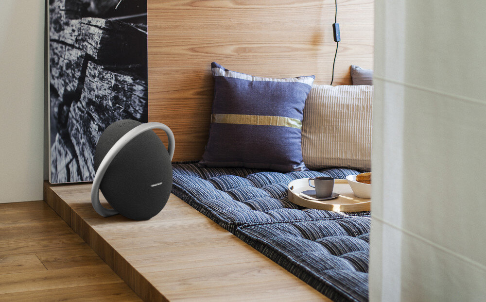 Czarny głośnik na drewnianej powierzchni. Poduszki i taca z jedzeniem w tle. Przytulne otoczenie.