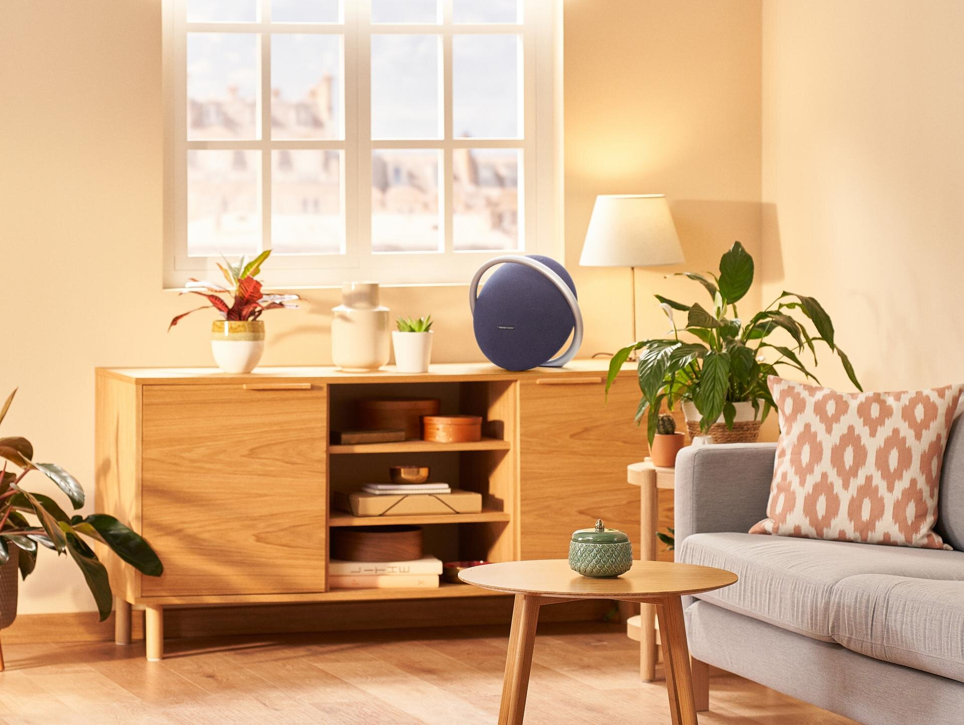 Salon z głośnikiem, drewnianą szafką, roślinami i oknem, przez które wpada światło słoneczne.