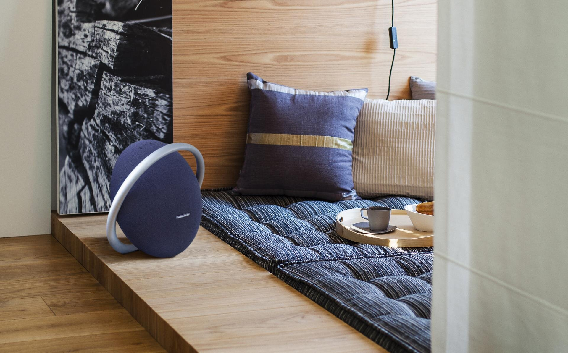 Niebieski głośnik na drewnianej platformie. Poduszki i taca z jedzeniem w tle.