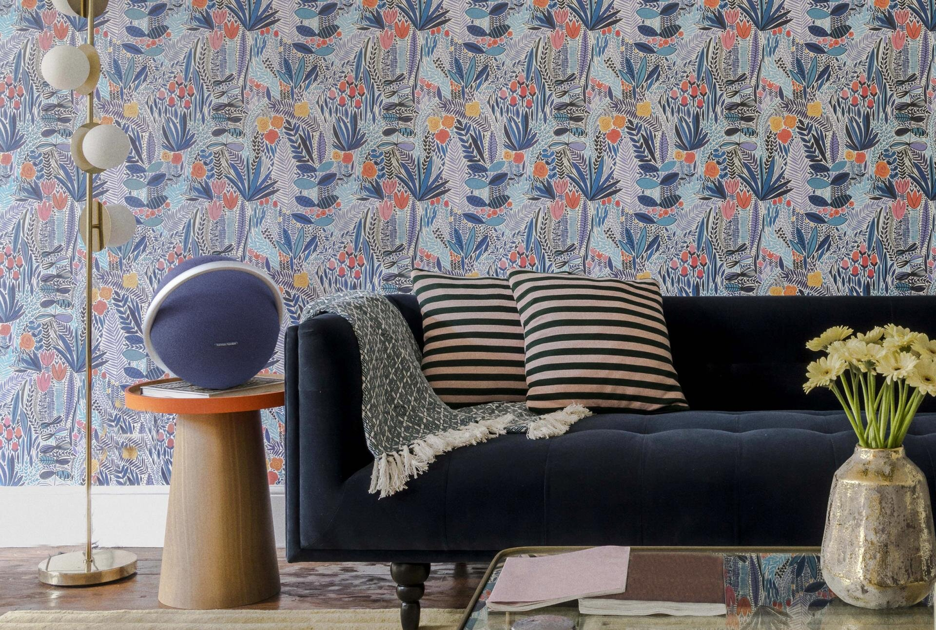 Niebieski głośnik na drewnianym stoliku, przed sofą i kwiatową tapetą.