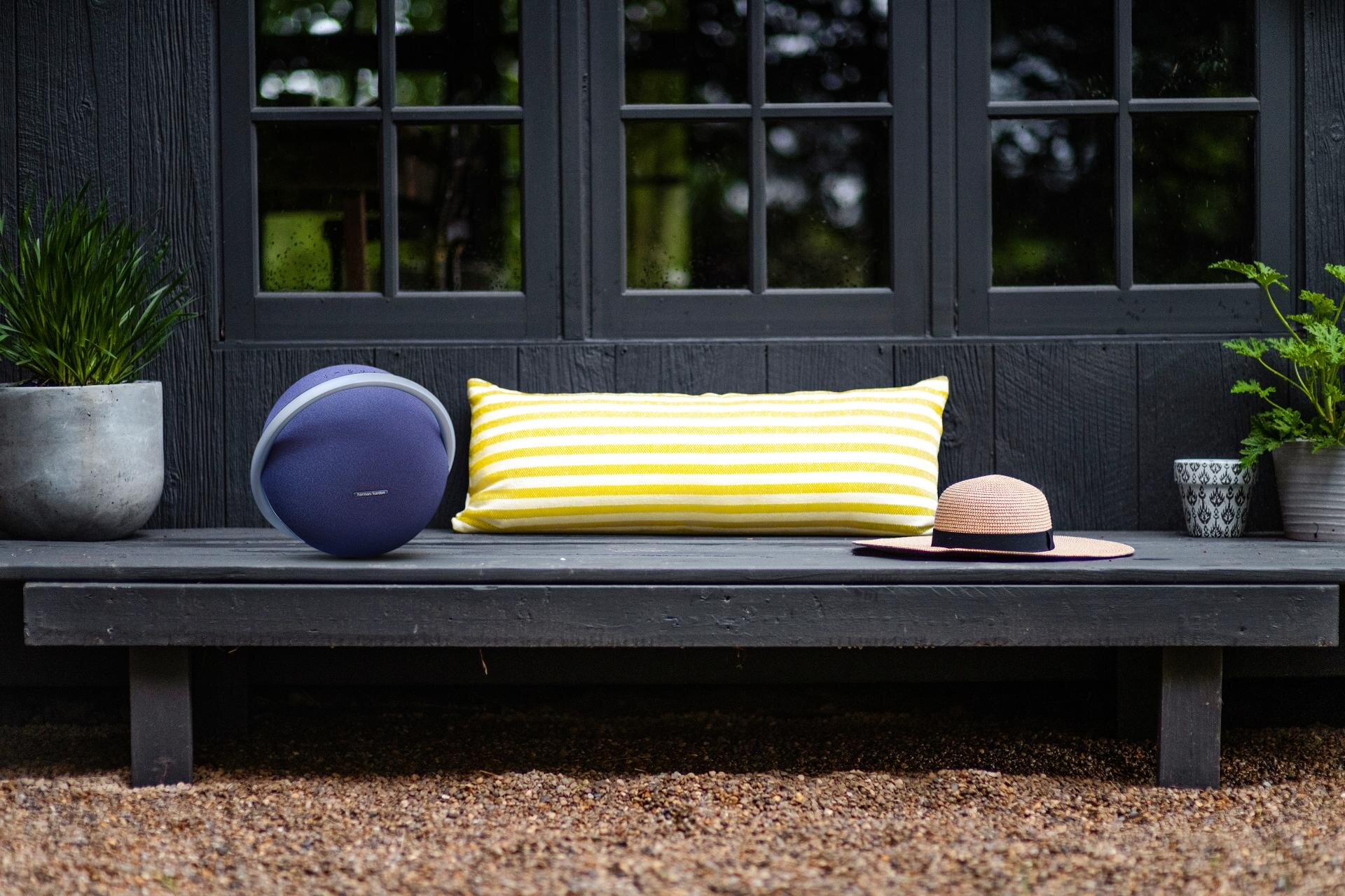 Scena na zewnątrz z niebieskim głośnikiem, żółtą poduszką w paski i kapeluszem na ciemnej werandzie.