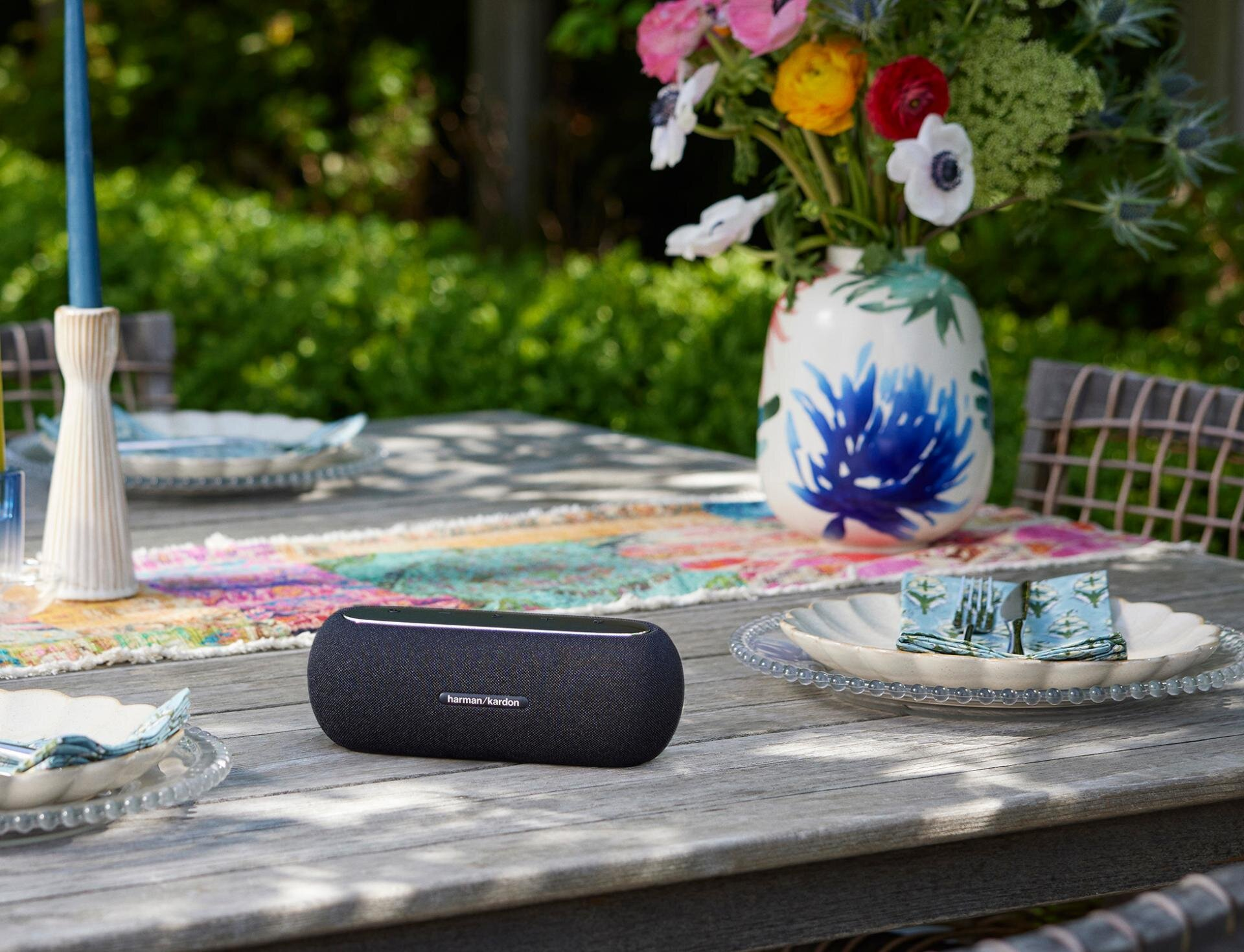 Czarny głośnik na drewnianym stole z kwiatami w tle. Obecne są talerze i kwiecisty bieżnik.