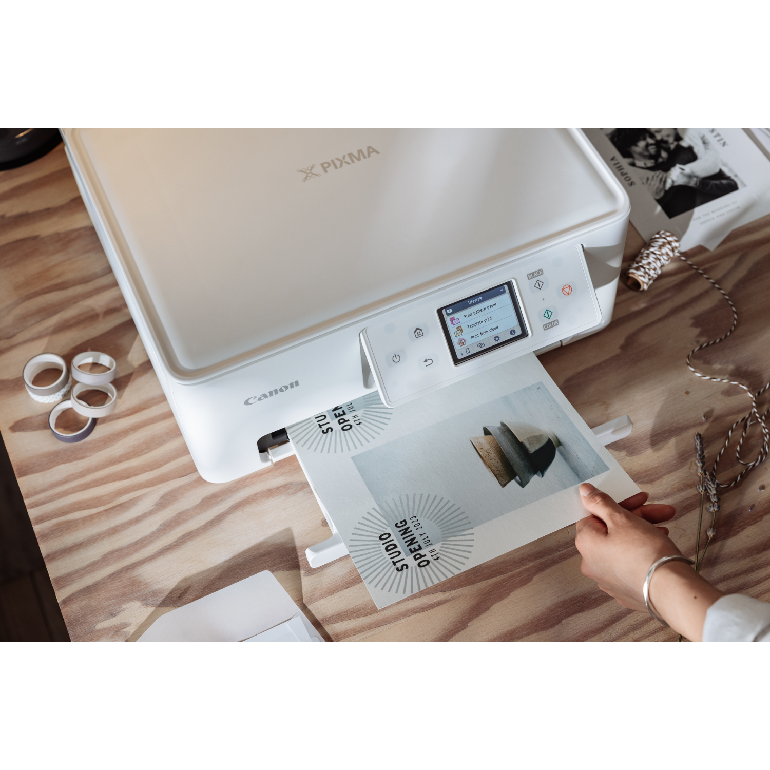 Een witte Canon Pixma printer die een brochure afdrukt. Een hand houdt een afgedrukt papier vast. Andere items liggen op de tafel.